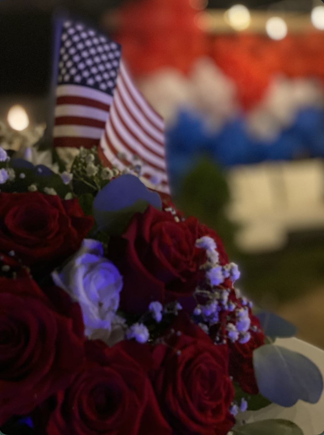 A floral arrangement of red roses with white baby's breath, an American flag, and a blurred patriotic cake in the background.