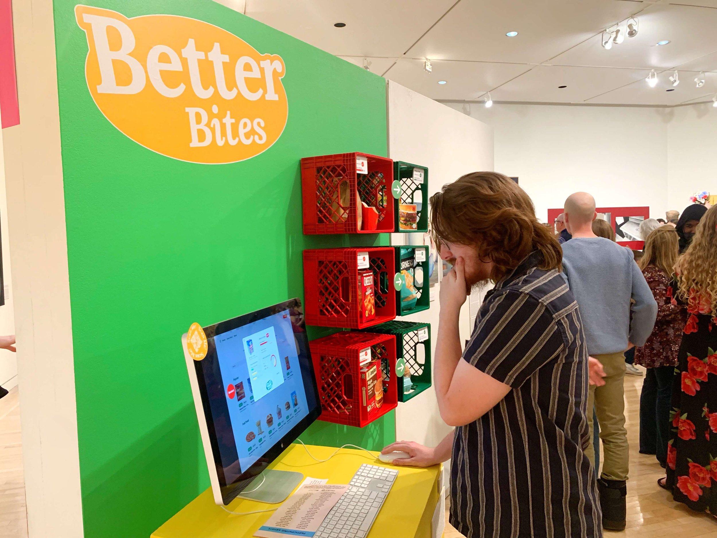 A woman with brown hair in a striped shirt is looking at a touchscreen ordering kiosk at Better Bites, a snack or food stand, with red and green shelves containing snack boxes behind her. Several people are in line in the background.