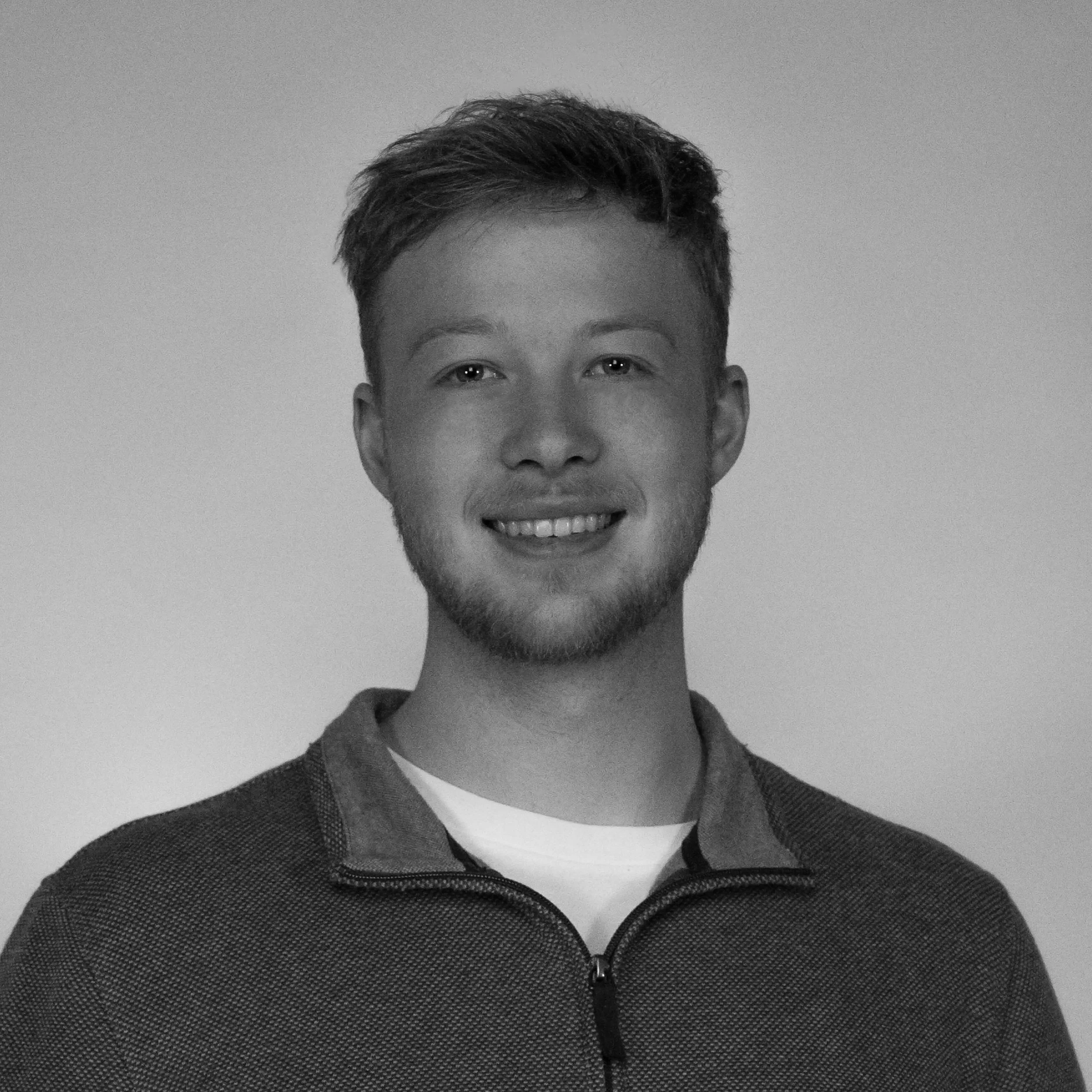 Black and white portrait of a smiling young man with short hair, a beard, wearing a zip-up sweater over a white shirt, standing against a plain background.