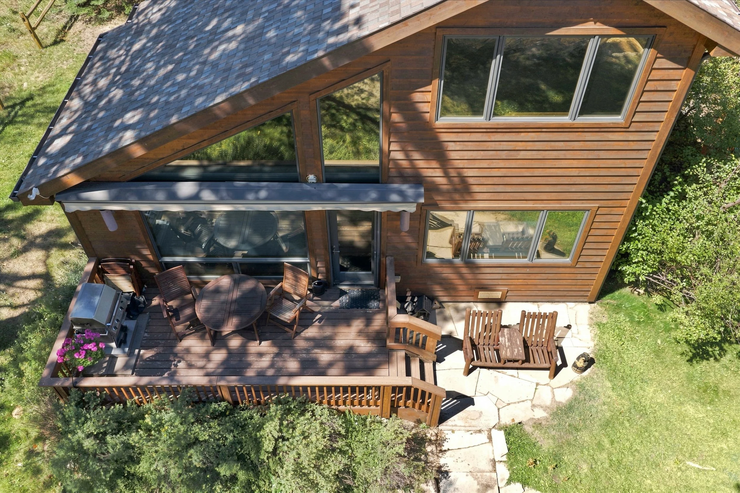 Top-down view of a wooden house with large windows and a deck. The deck has a round dining table with six chairs, an outdoor grill, a small potted plant with pink flowers, and a wood privacy screen. Surrounding the house are green trees and grass.