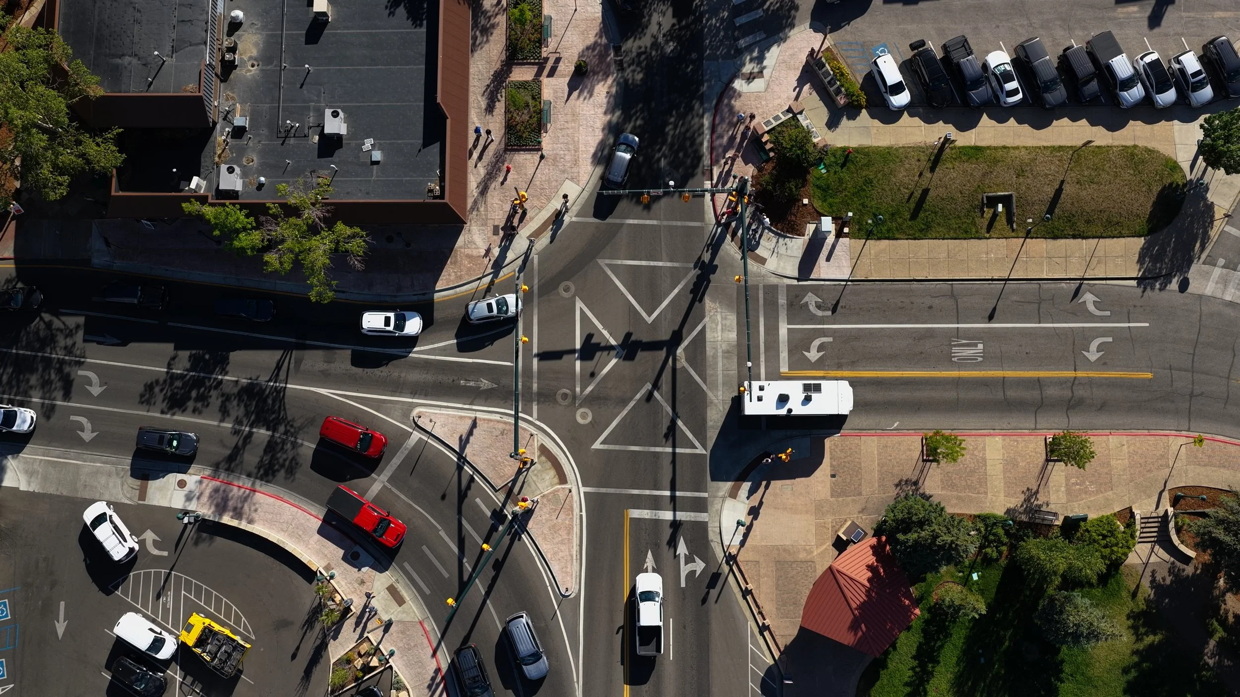 An aerial view of a city street intersection with cars, parking lots, trees, and pedestrian sidewalks, showing road markings and crosswalks