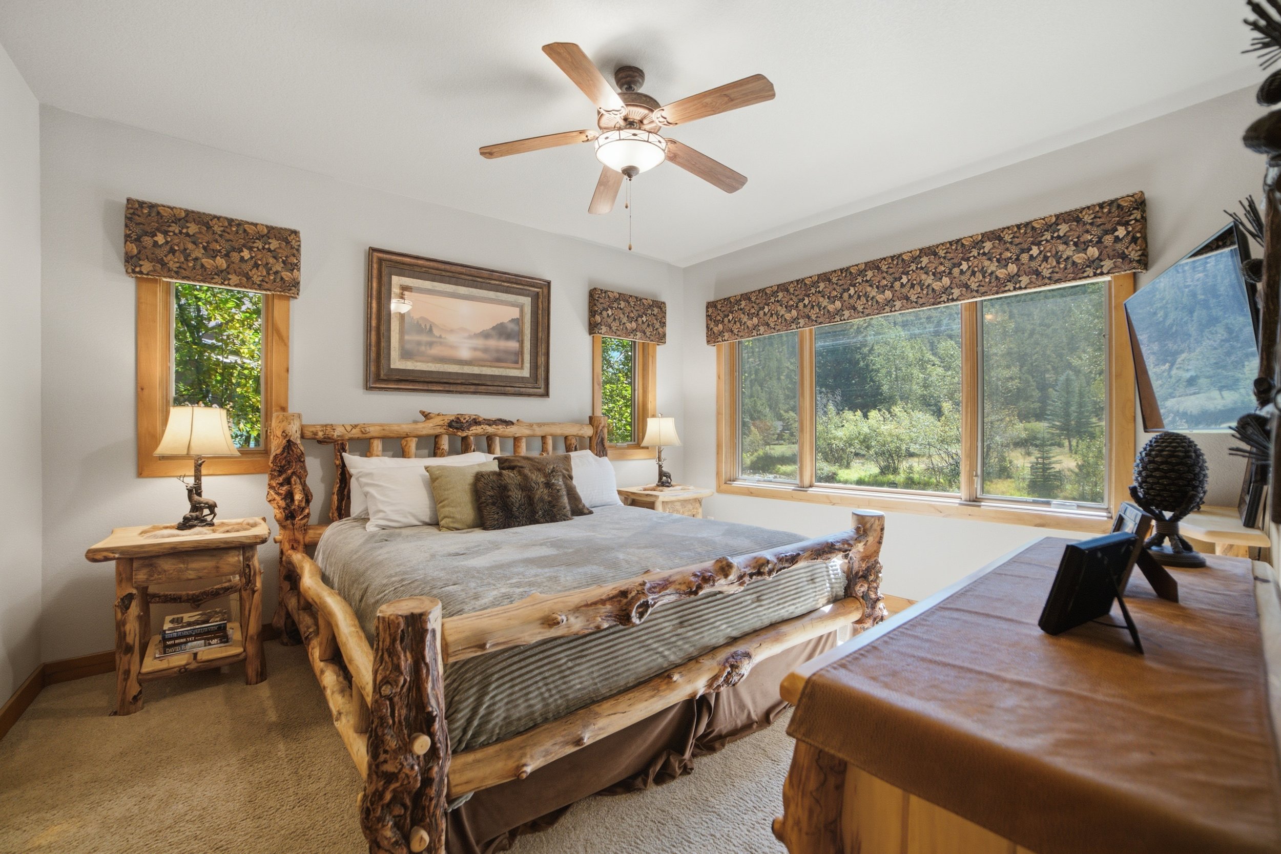 A rustic bedroom with a log bed frame, a nightstand with a lamp, and large windows with floral valances showing green trees outside.