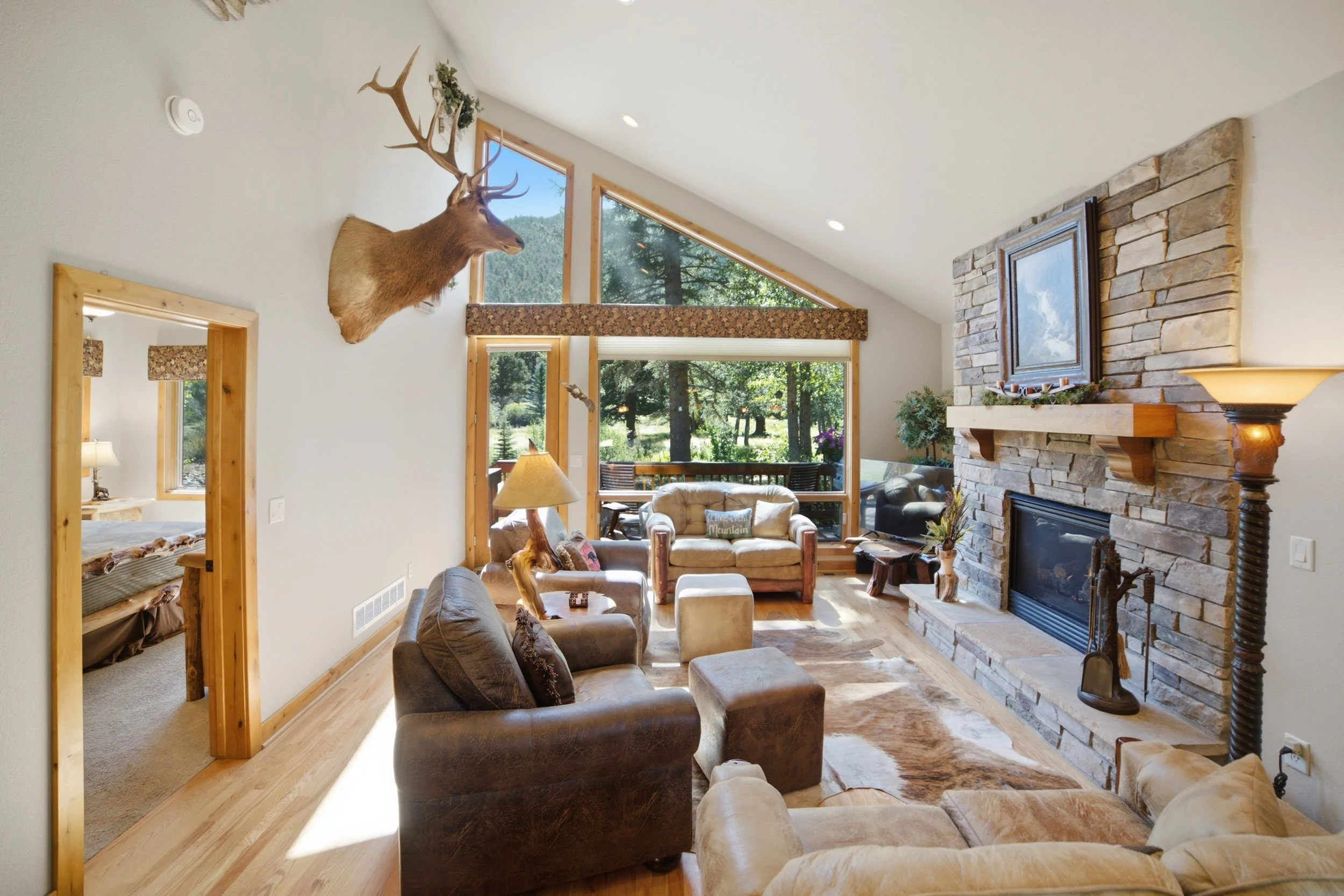 A cozy living room with large windows showing trees outside, a stone fireplace with a framed picture above, a mounted deer head on the wall, and comfortable seating.