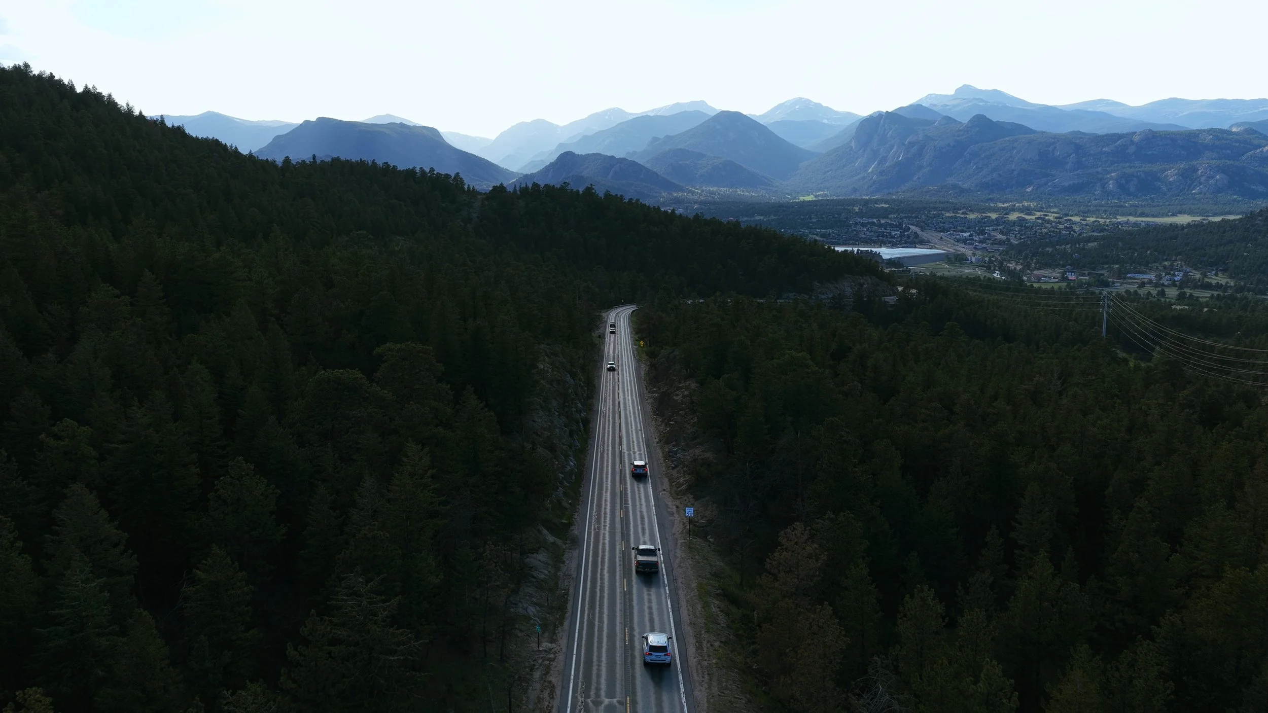 A mountain landscape with a valley, distant mountain ranges with snowy peaks, and a highway with cars traveling through a forested area.
