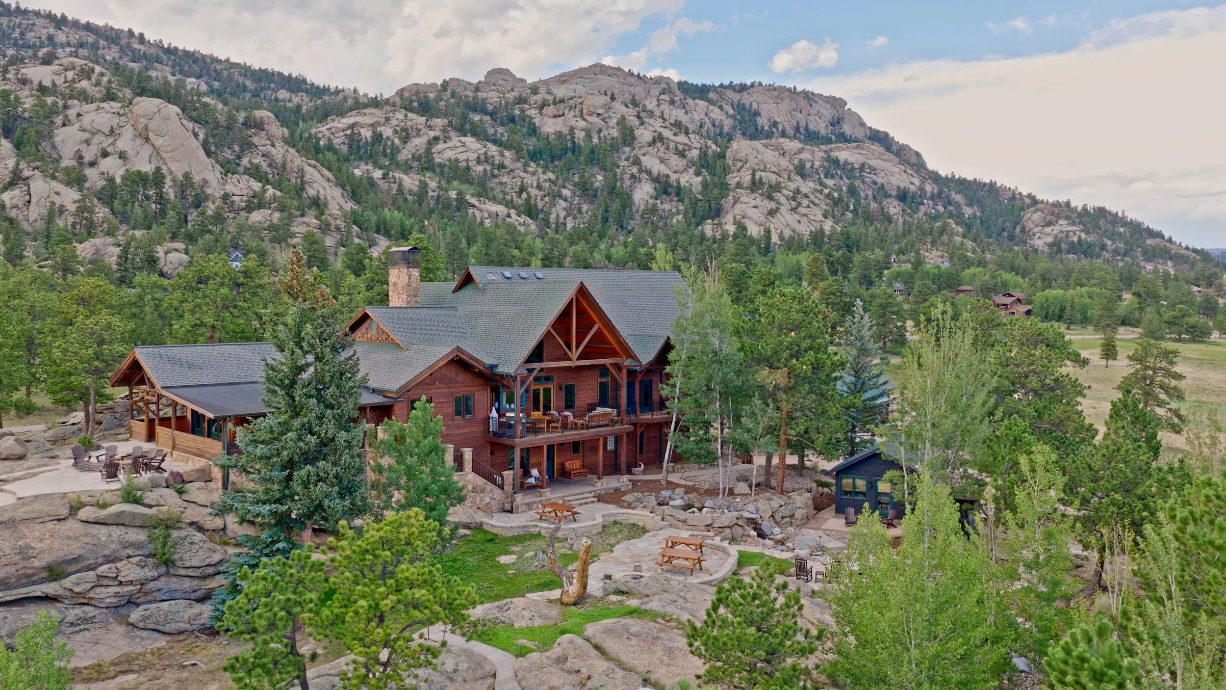 A large wooden house in a mountainous area surrounded by trees and rocky terrain, with a patio and outdoor seating.