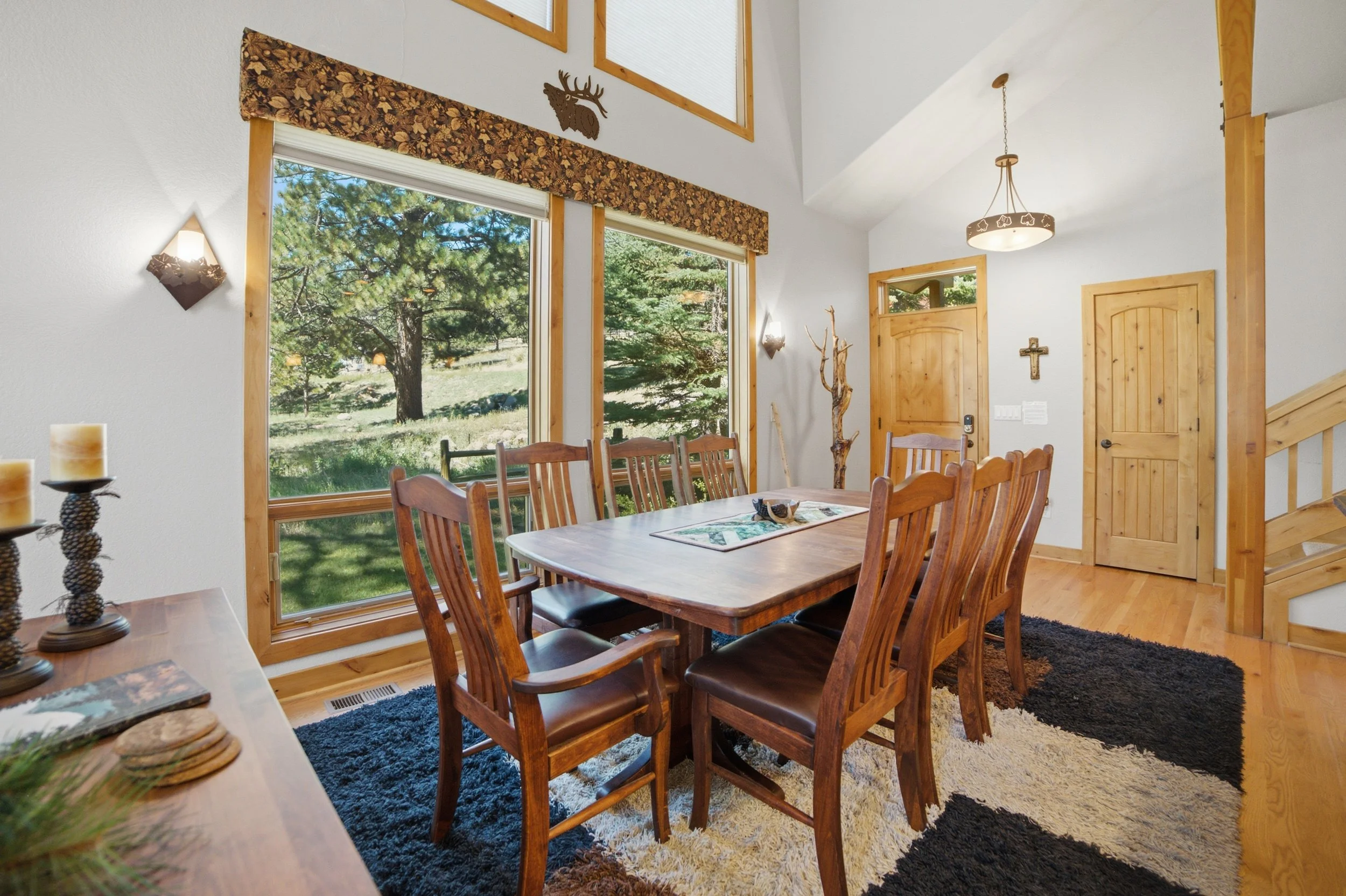 A dining area inside a log cabin with large windows showing a forested landscape outside. The room features a wooden table with several matching chairs, wall-mounted light fixtures, and decorative items including a wall cross and a metal deer head silhouette. The space has wooden accents and a mix of dark and light-colored rugs on a hardwood floor.
