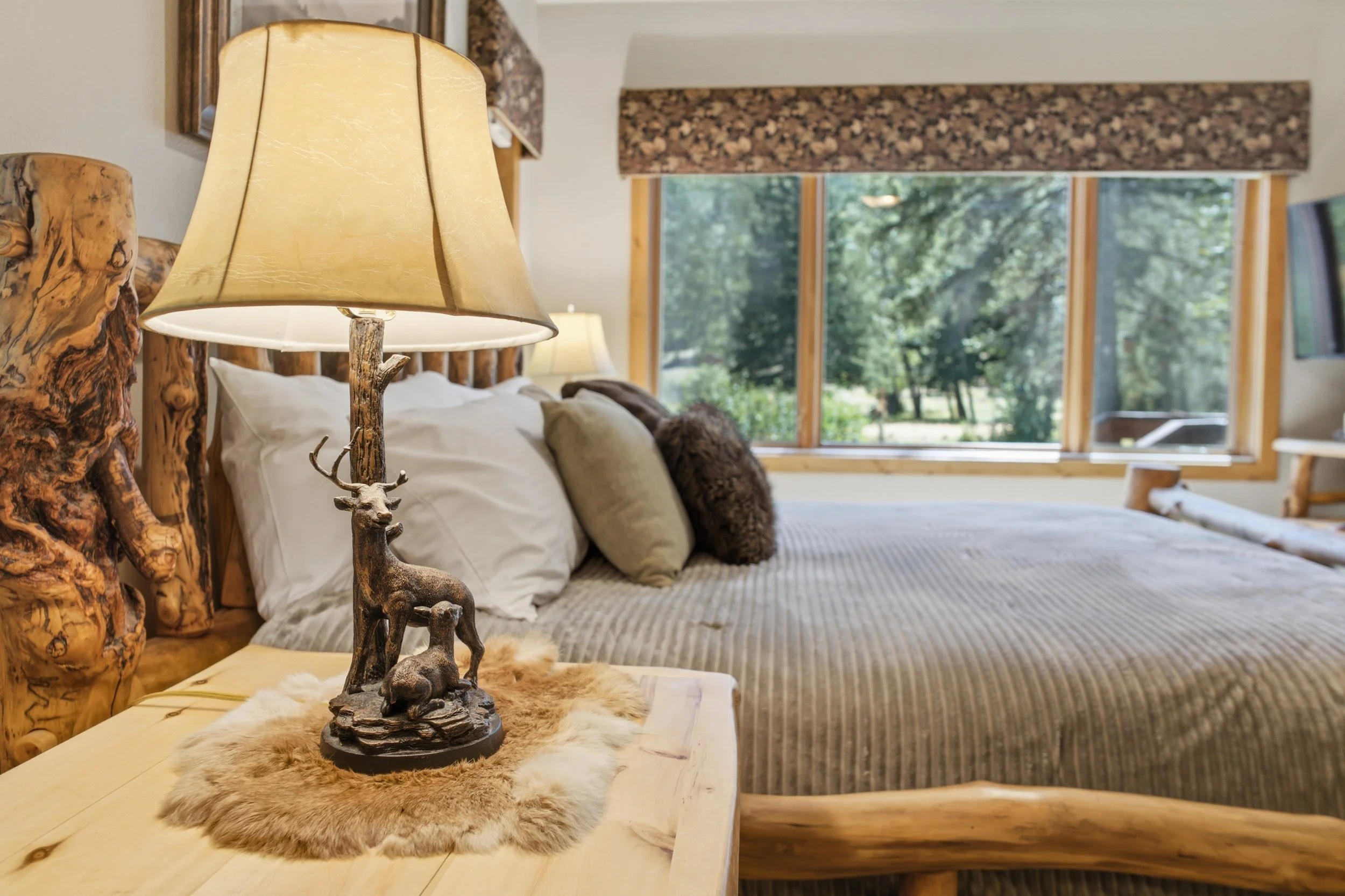 A rustic bedroom with a wooden bed frame and natural wood accents. A nightstand with a deer figurine and fur rug. Large windows with trees outside, letting in natural light.