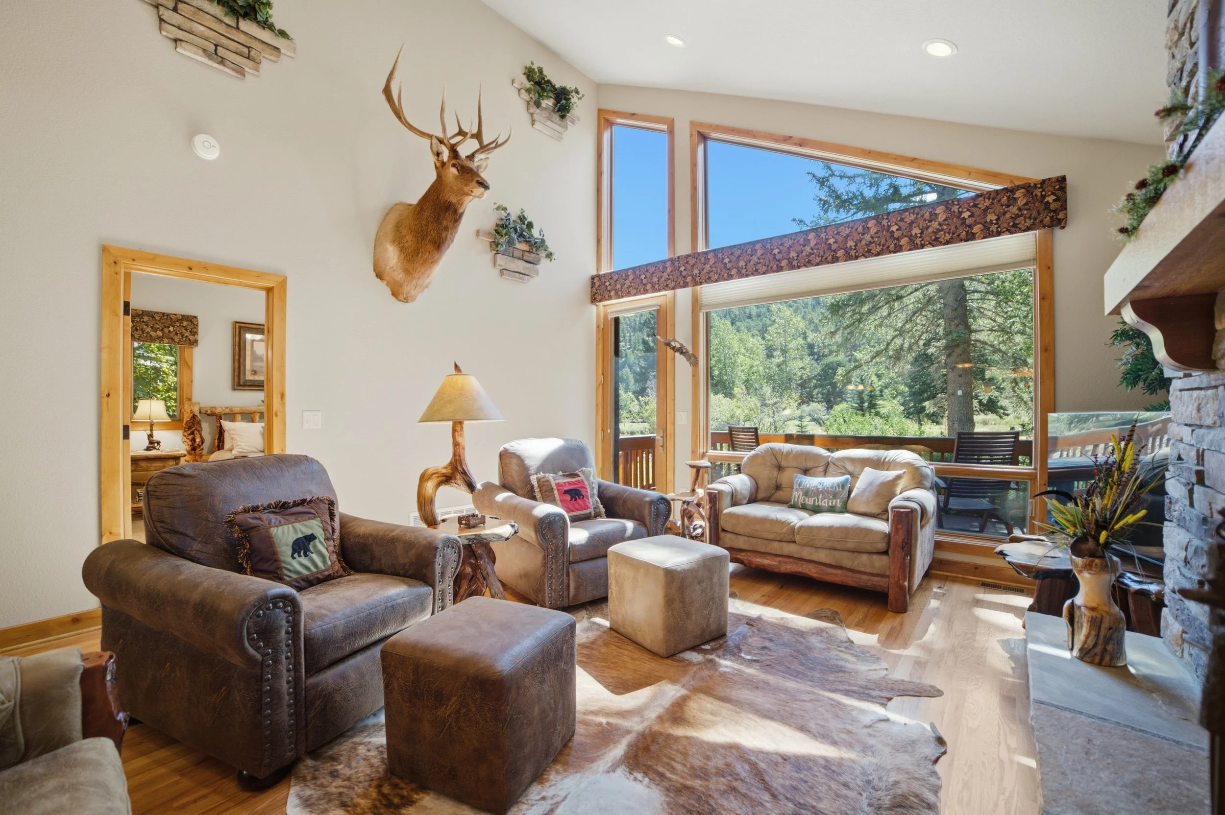 Living room with large windows showing trees outside, decorated with a mounted deer head, wall planters, and rustic furniture.