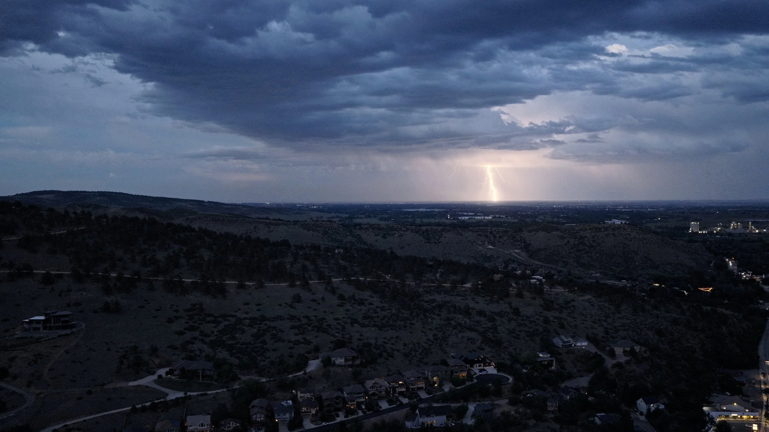 A dark, cloudy sky over a hilly residential area with houses and buildings. A lightning bolt illuminates the horizon in the distance during a thunderstorm.