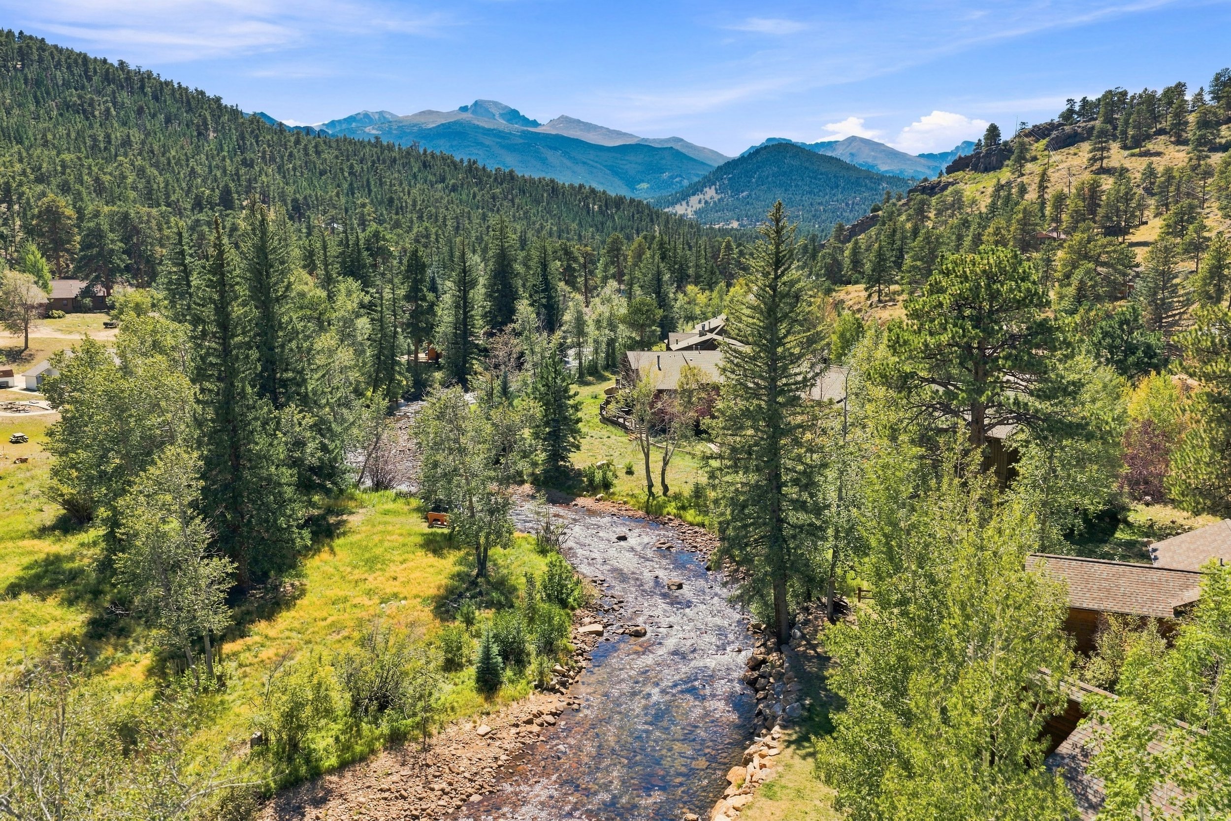 Scenic view of a mountain landscape with a river running through a forested valley, houses visible among the trees, and mountains in the distance under a blue sky.