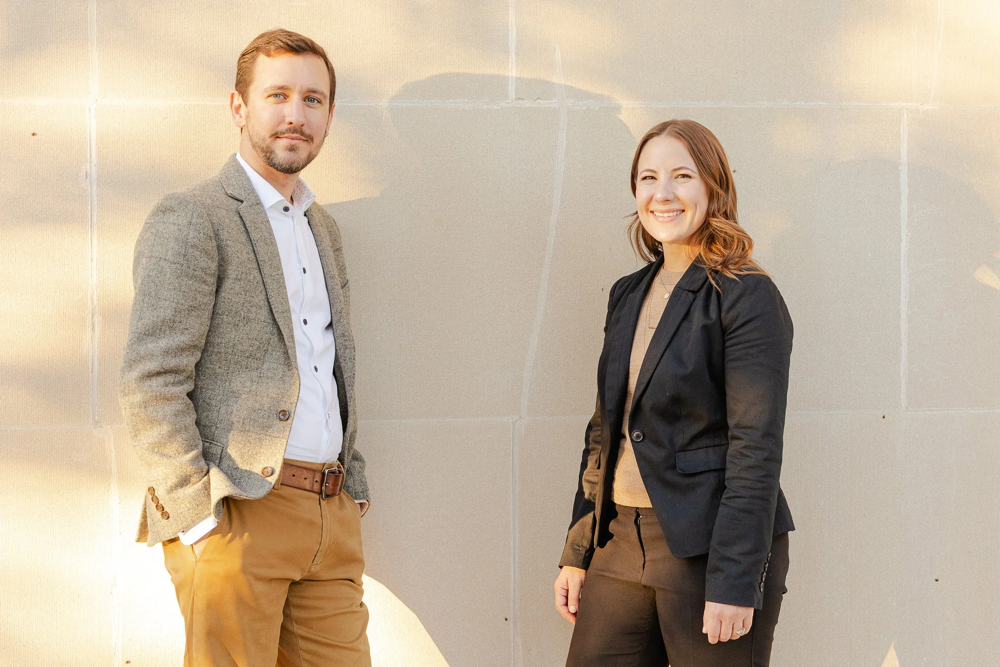 A man and a woman standing outdoors in front of a beige tiled wall, smiling and looking at the camera, with sunlight casting shadows.