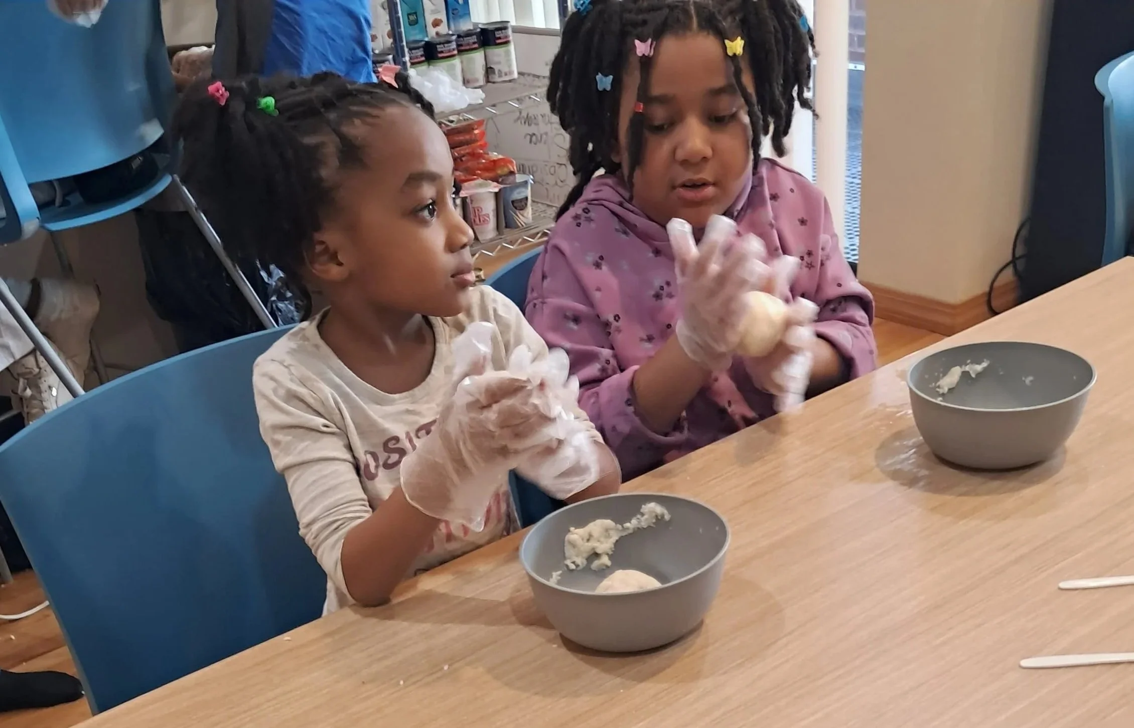 Two young girls with braided hair and colorful butterfly clips, wearing gloves, sitting at a table with bowls of dough, in a room that appears to be a bakery or kitchen environment.