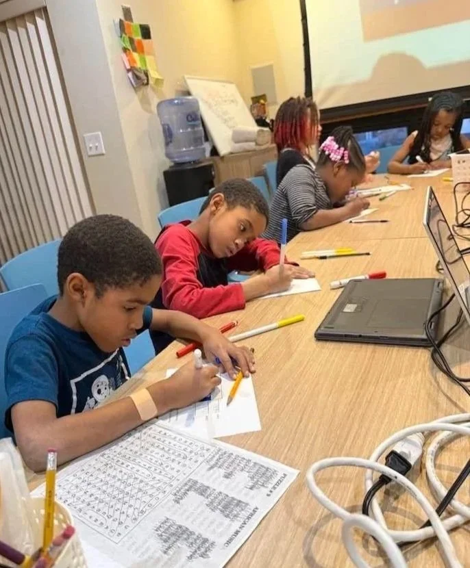 Children sitting at a table in a classroom, working on worksheets with pens and pencils, with a water dispenser and a screen in the background.