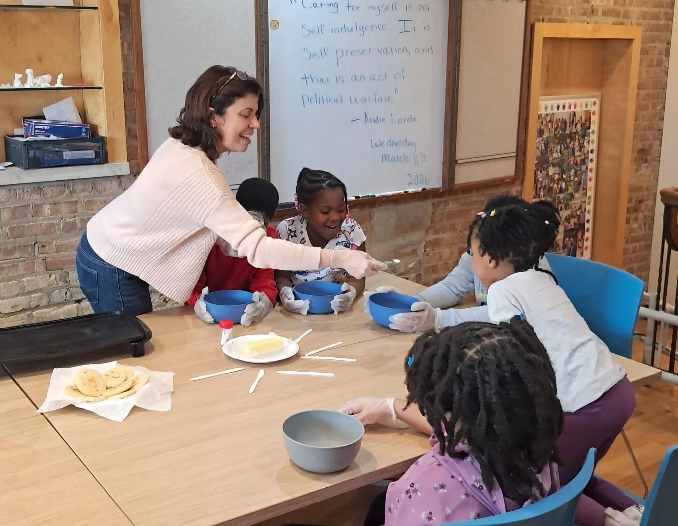 A woman and four children seated around a table in a classroom, engaging in a craft activity with bowls. The background shows a brick wall, a whiteboard with writing, and shelves with books and decor.