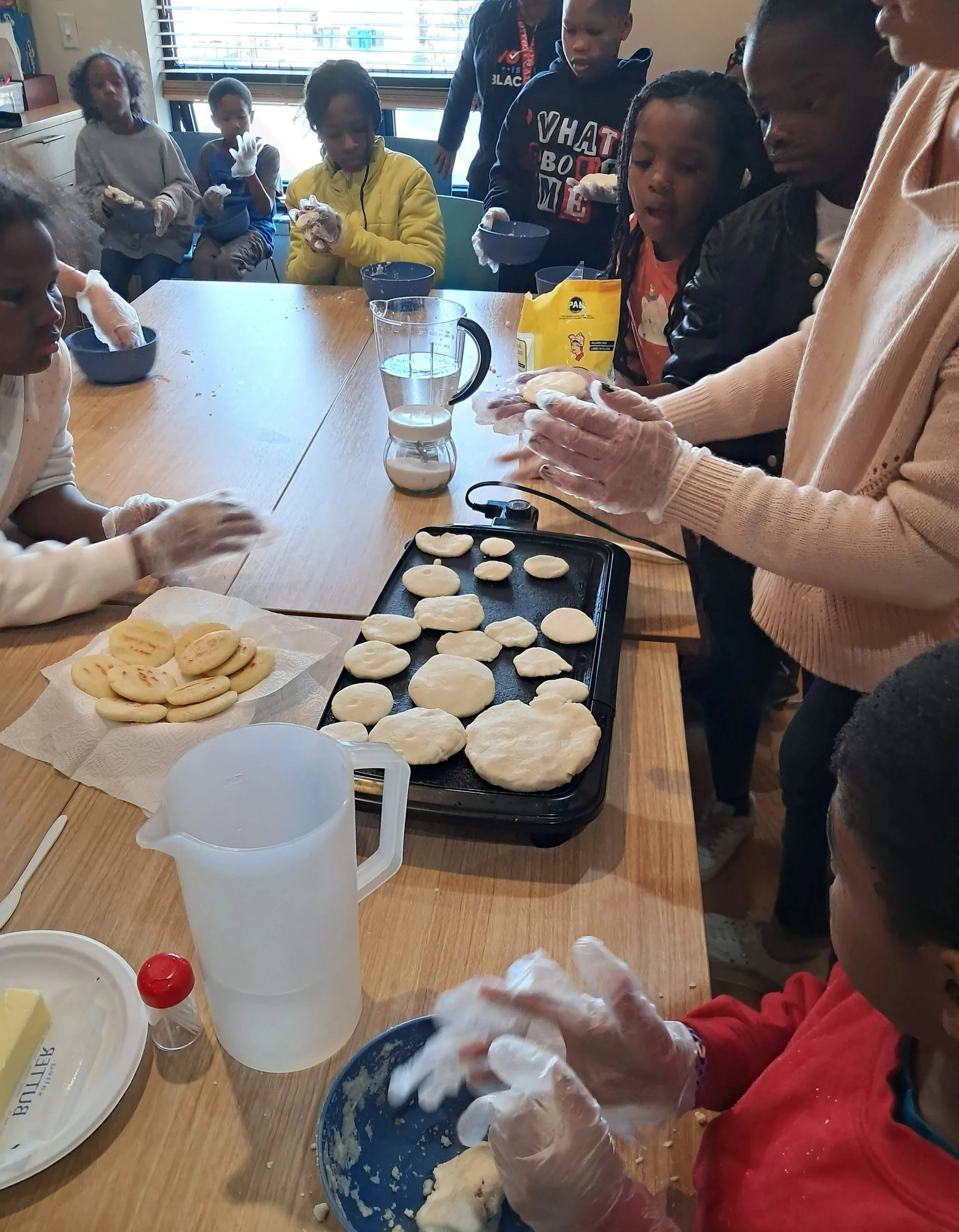 Children and adults participating in a baking activity, kneading dough at a table, with baking ingredients and tools around, in a room with windows and blinds.