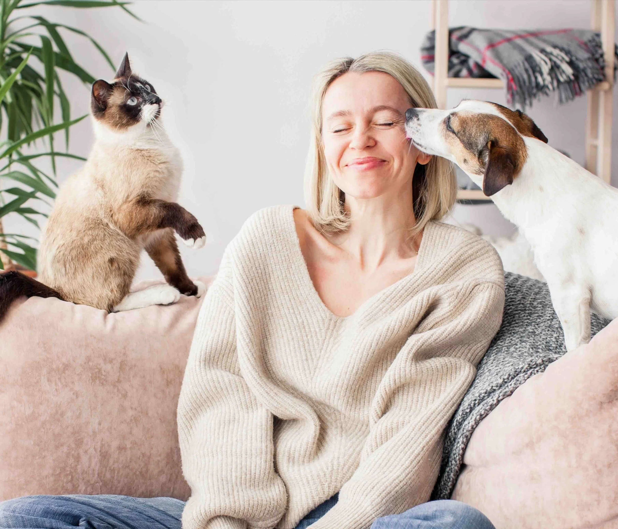 A woman sitting on a couch with a Siamese cat on her left and a dog on her right, both reaching out to her face, in a cozy living room.