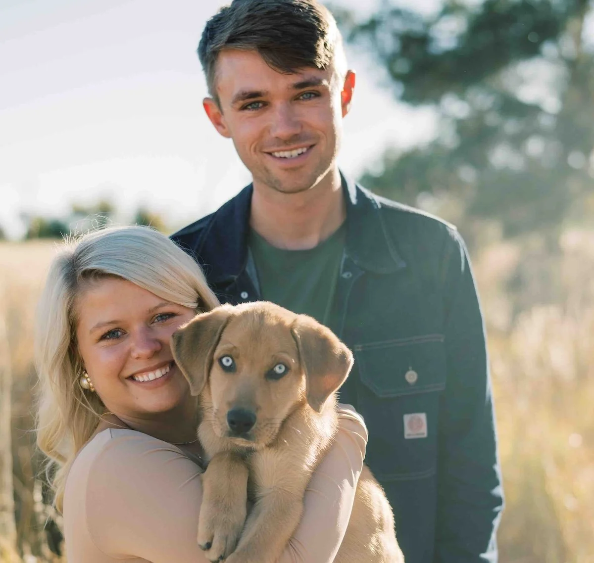 A smiling woman holding a puppy with blue eyes, standing outdoors with a man in the background.