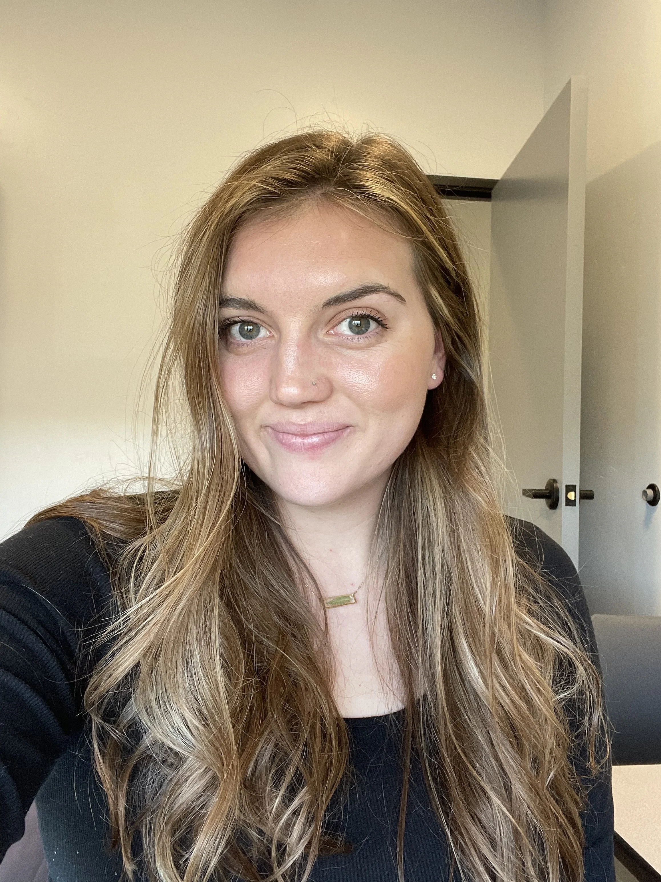 A woman with long wavy light brown hair, fair skin, and blue eyes, smiling and wearing a black top and a gold rectangular necklace, taking a selfie in a room with light-colored walls and a closed door in the background.