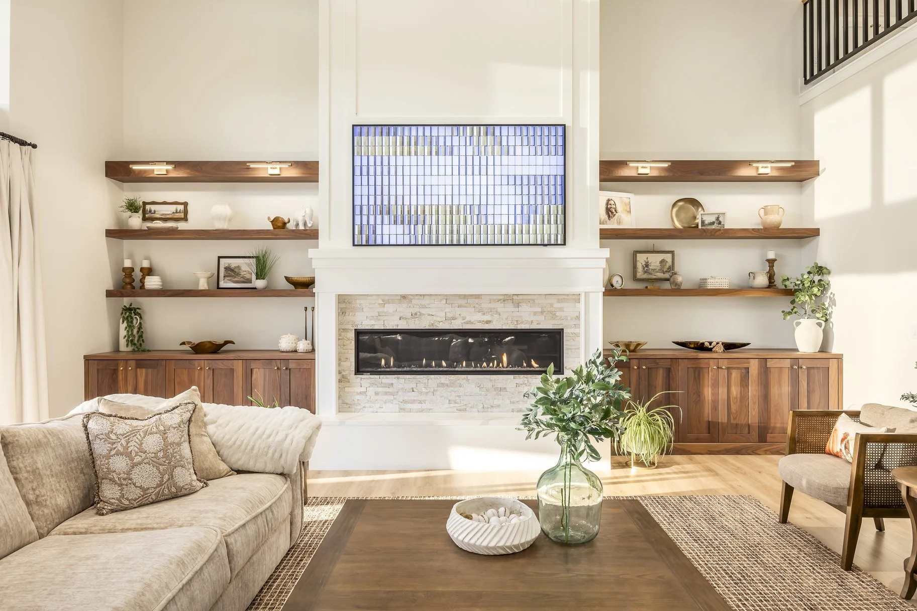 Living room with fireplace, TV, wooden shelves with decorative items, beige sofa, plants, and natural light.