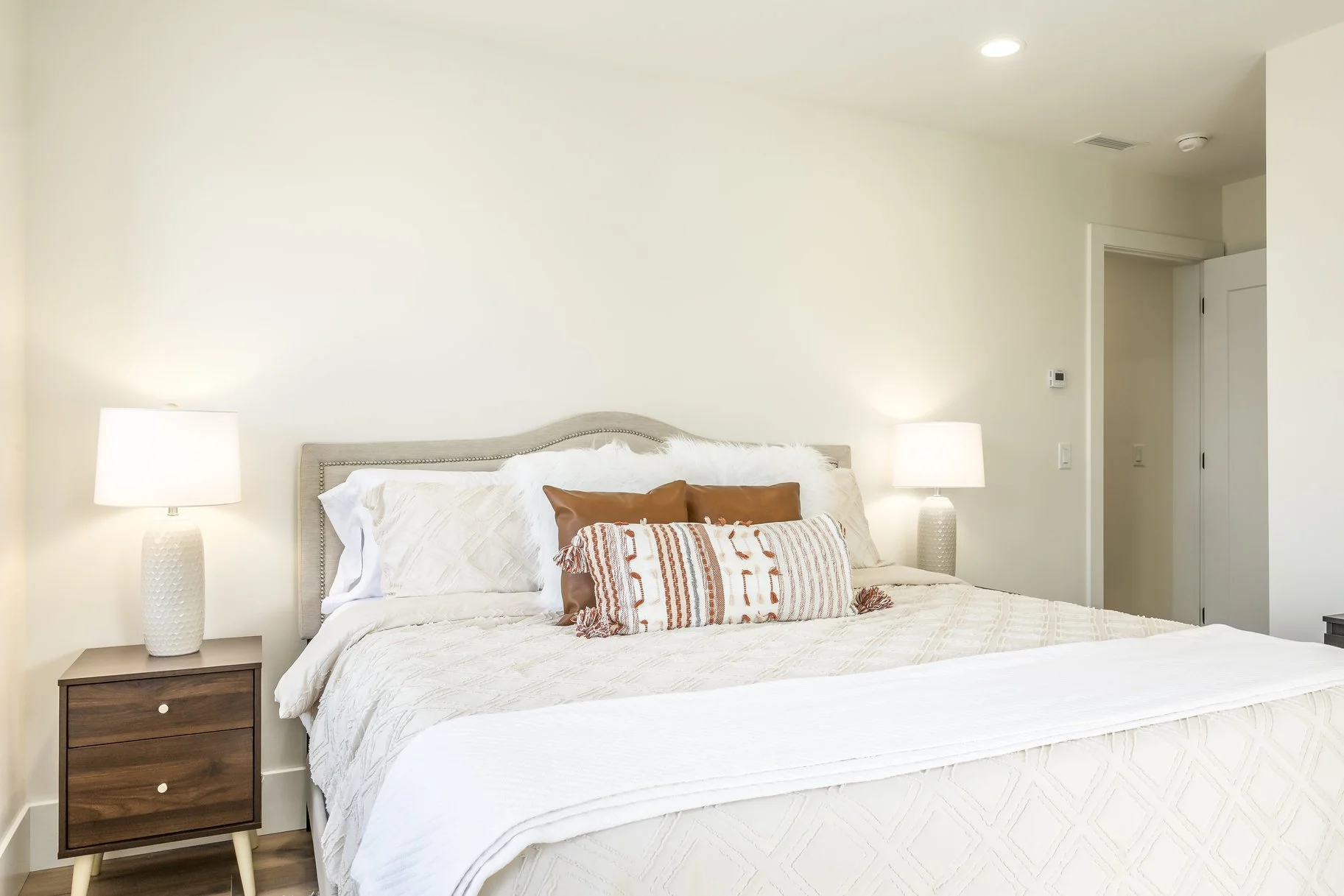 A neatly made bed with white bedding, decorated with assorted pillows, in a bedroom with nightstands and lamps on each side.
