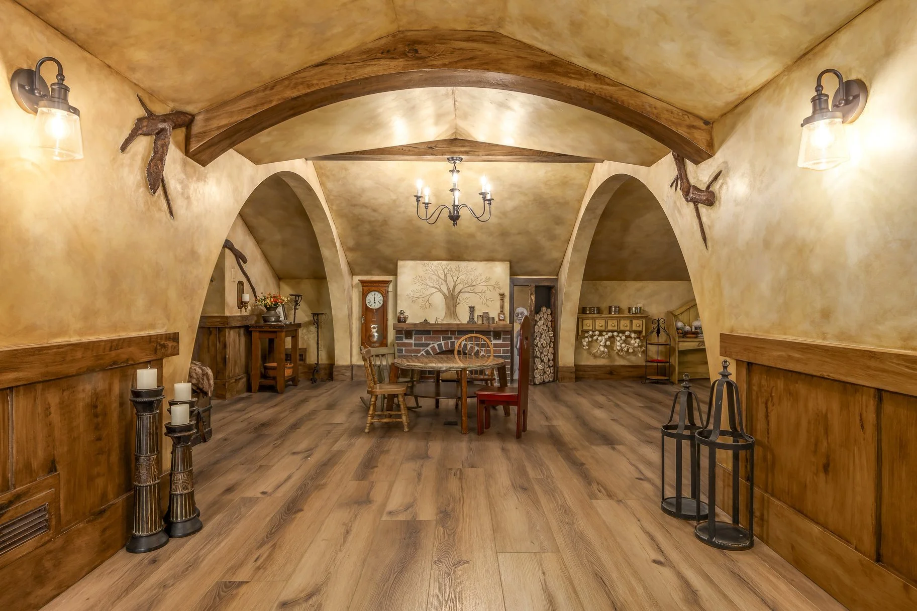 A cozy dining room with arched ceilings, wooden beams, and warm yellow walls. It features a round wooden table with three chairs, a grandfather clock, and a brick fireplace with a painting of a tree above it. Decorative items include candles, a wall-