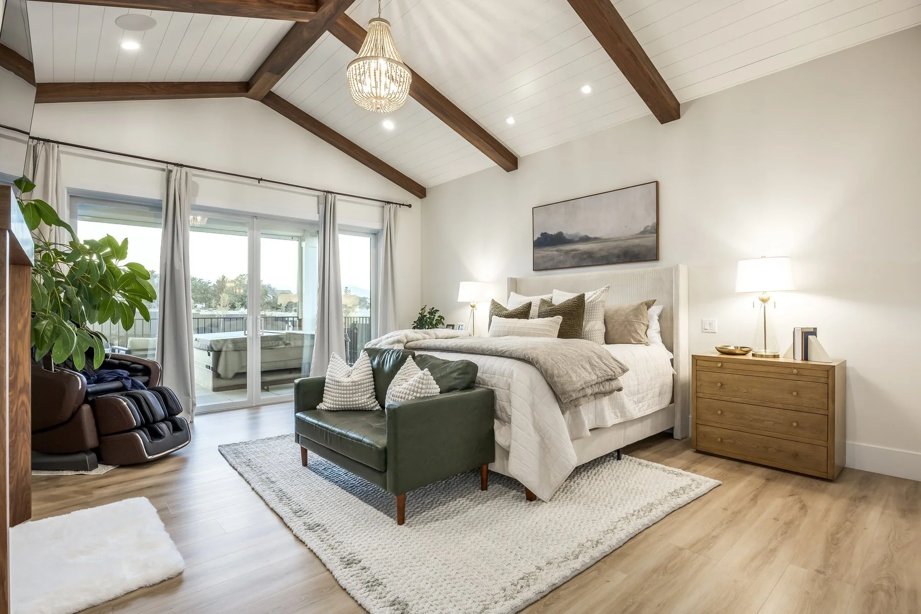 A bedroom with a vaulted ceiling, large sliding glass door leading to a balcony, a bed with white linens and multiple pillows, a wooden nightstand with a lamp, a green armchair at the foot of the bed, and a massage chair to the left near the sliding 