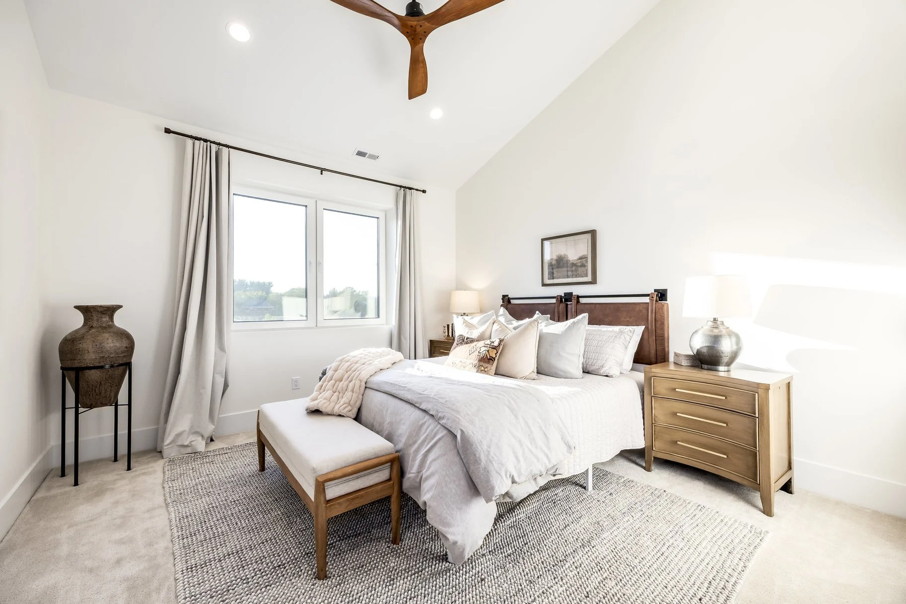 A bright, cozy bedroom with a large bed, wooden headboard, various pillows, a nightstand with a lamp, a window with curtains, a decorative vase on a stand, and a textured beige rug.