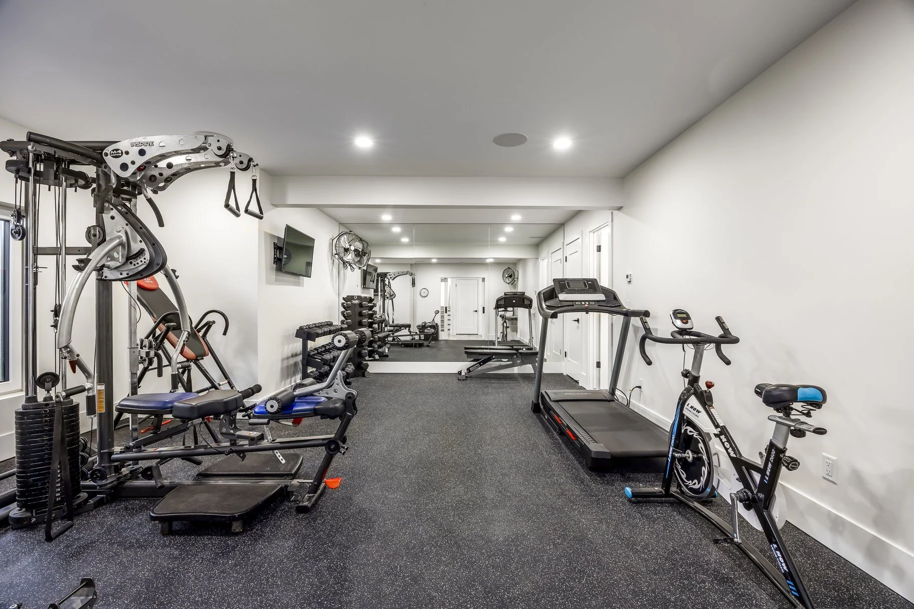 Clean home gym with various exercise equipment including a treadmill, stationary bike, weight machine, free weights, and a TV on the wall.