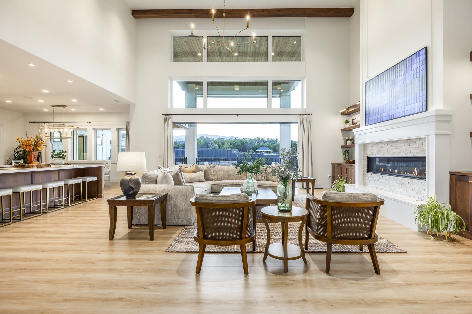 Bright open-concept living room with high ceiling, large windows, beige sectional sofa, wooden armchairs, fireplace, and modern lighting fixtures.