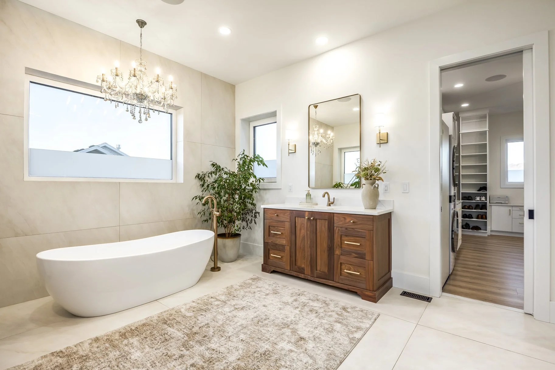 Modern bathroom with a freestanding bathtub, wooden vanity with a white countertop, large window, chandelier, mirror, potted plant, and laundry room visible through an open door.