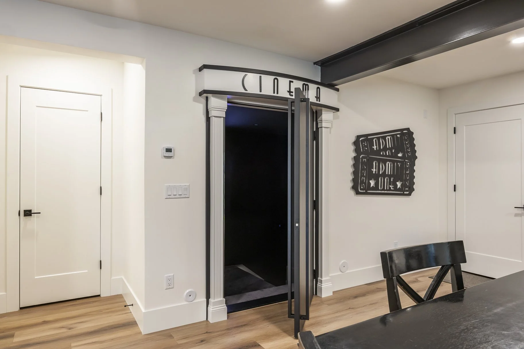 Interior view of a modern home, featuring a black elevator with its doors open, decorative signs on the wall, a white door, and a wooden dining table with black chairs.