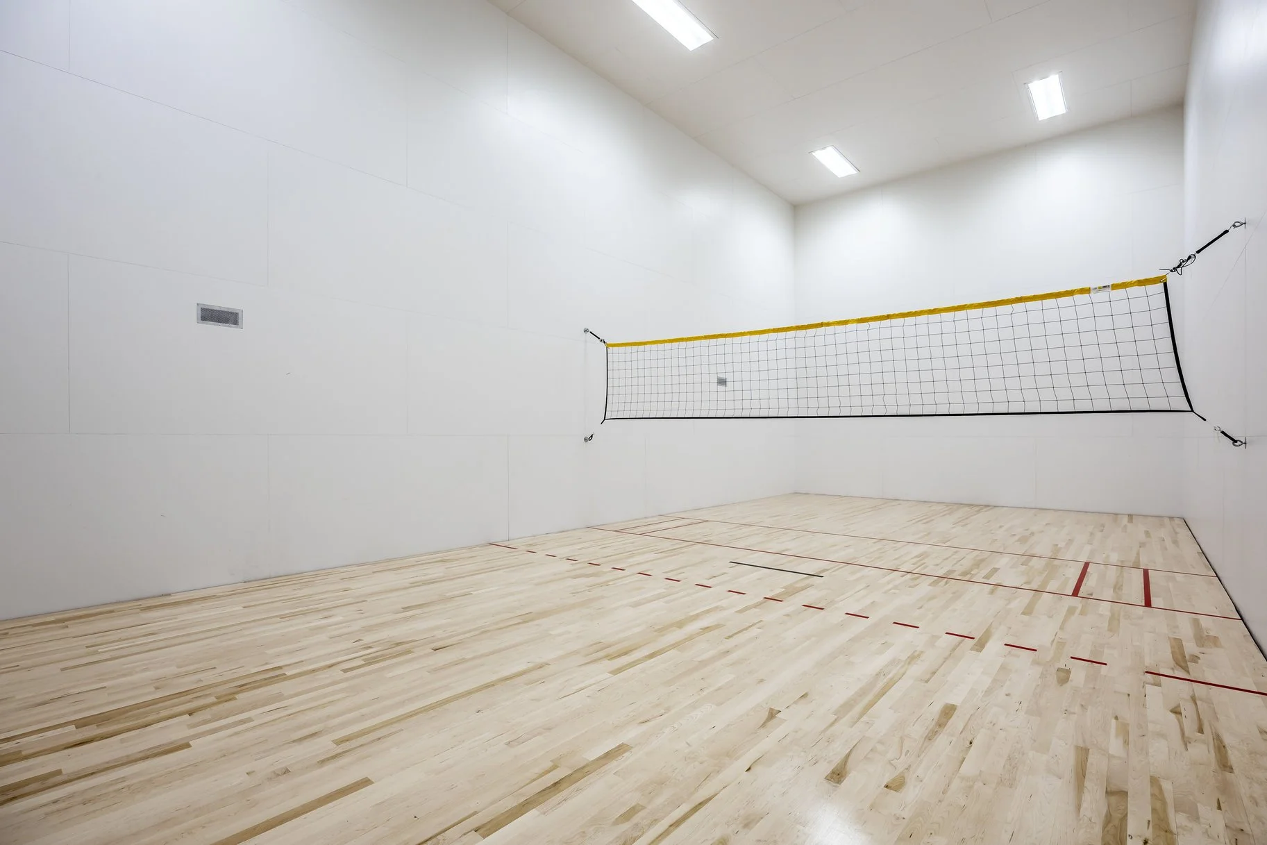 An indoor volleyball court with light-colored wooden flooring, white walls, and a volleyball net set up in the middle.