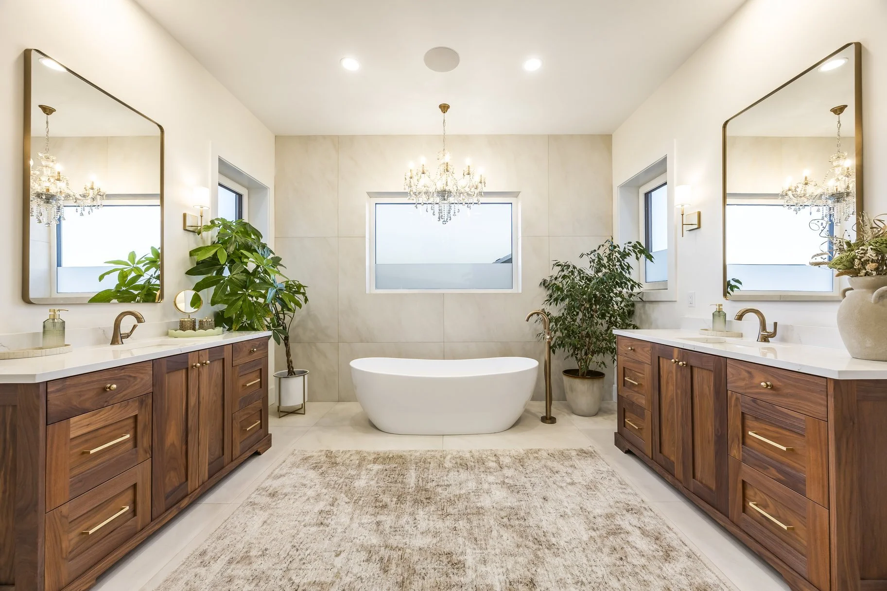 A luxurious bathroom with dual wooden vanities, large mirrors, a freestanding bathtub, potted plants, and a chandelier.