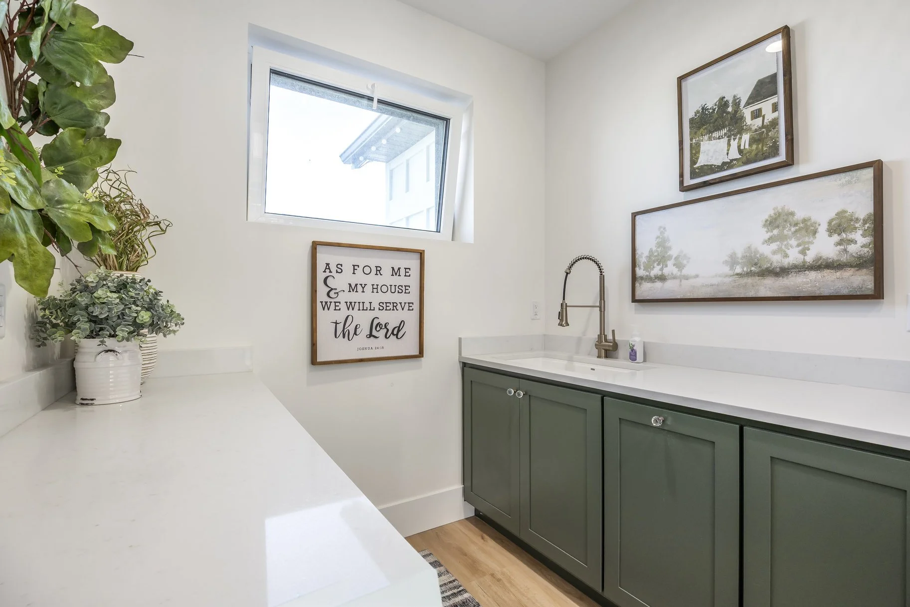 A laundry or utility room with a white countertop, green cabinets, and a window. There are framed wall art, including a biblical quote, and paintings of landscapes. A potted plant is on the countertop.