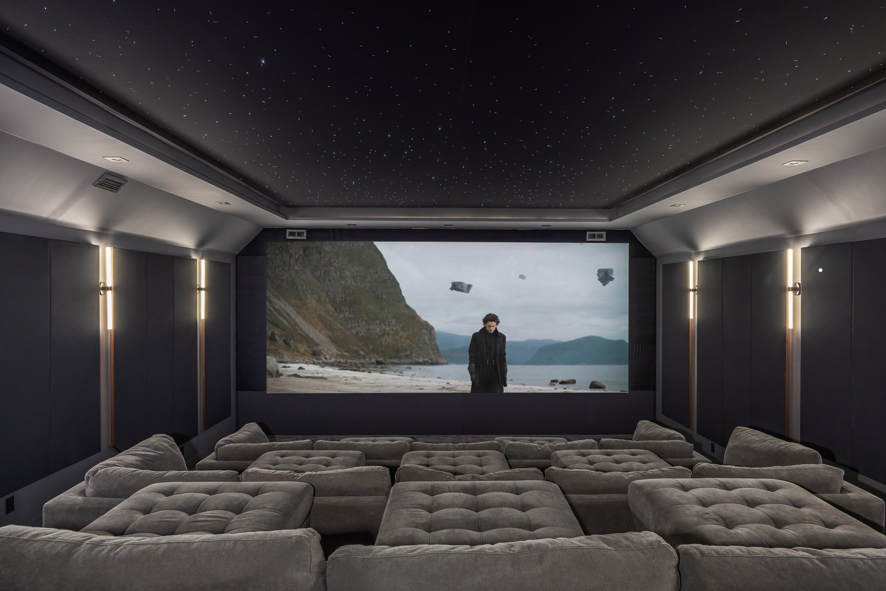 Modern home theater room with plush beige seating, dark walls, and a ceiling decorated with star-like lights, showing a scene from a movie with a person on a beach and floating debris.