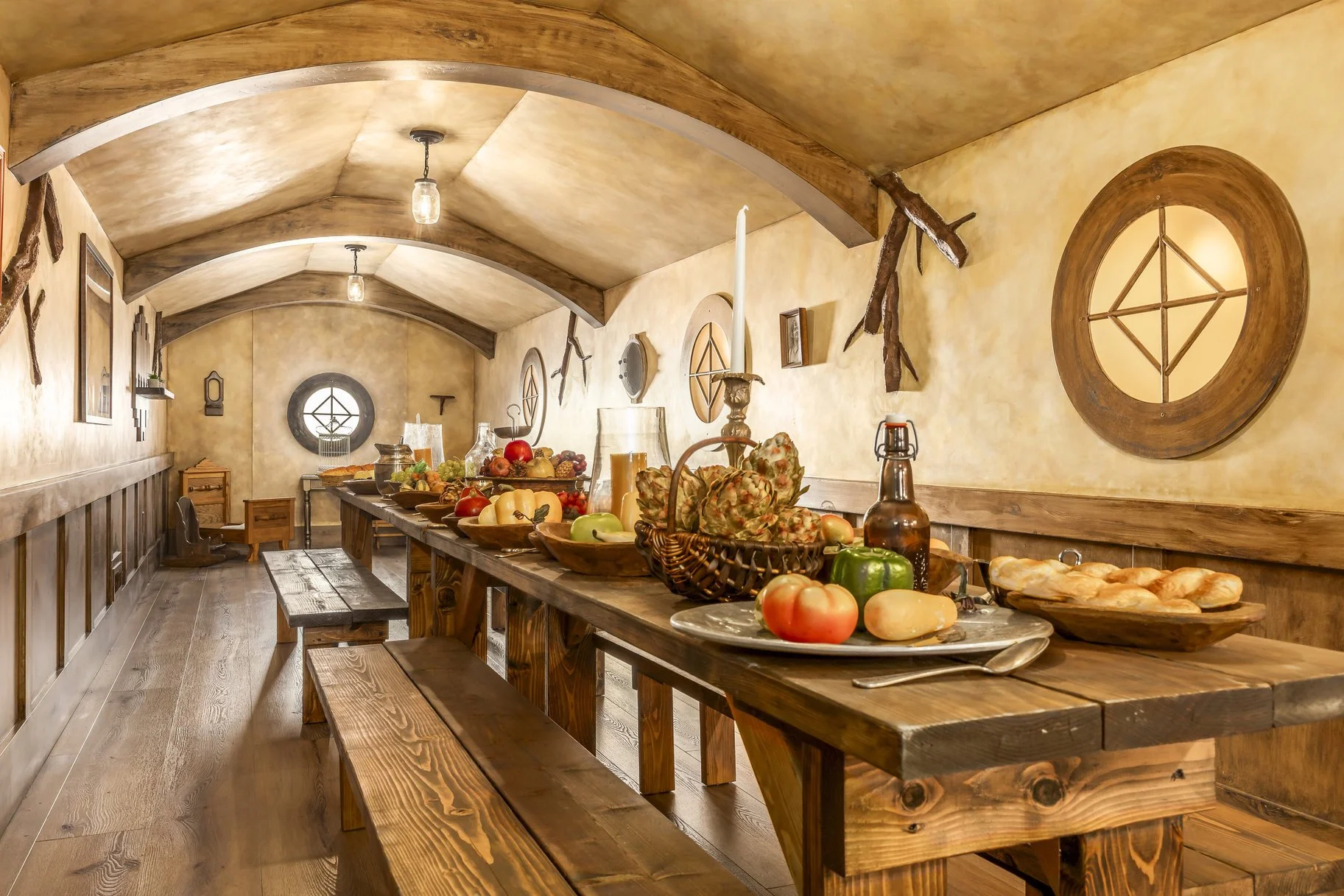 A rustic dining room with wooden tables and benches, decorated with fruits, vegetables, and candles, featuring walls with mounted antique tools and circular windows.