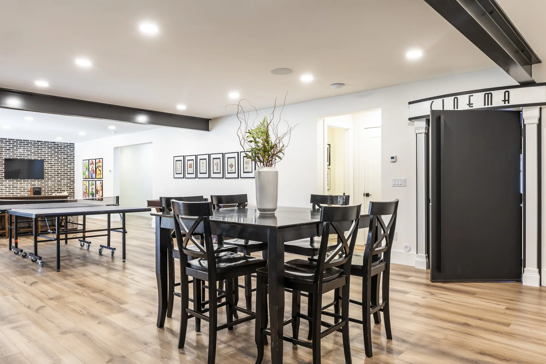 Modern indoor recreational room with a ping pong table, a round dining table with six black chairs, and a decorative vase with dried branches. There's a hallway and a black-and-white photo wall in the background, and a theater entrance to the right.