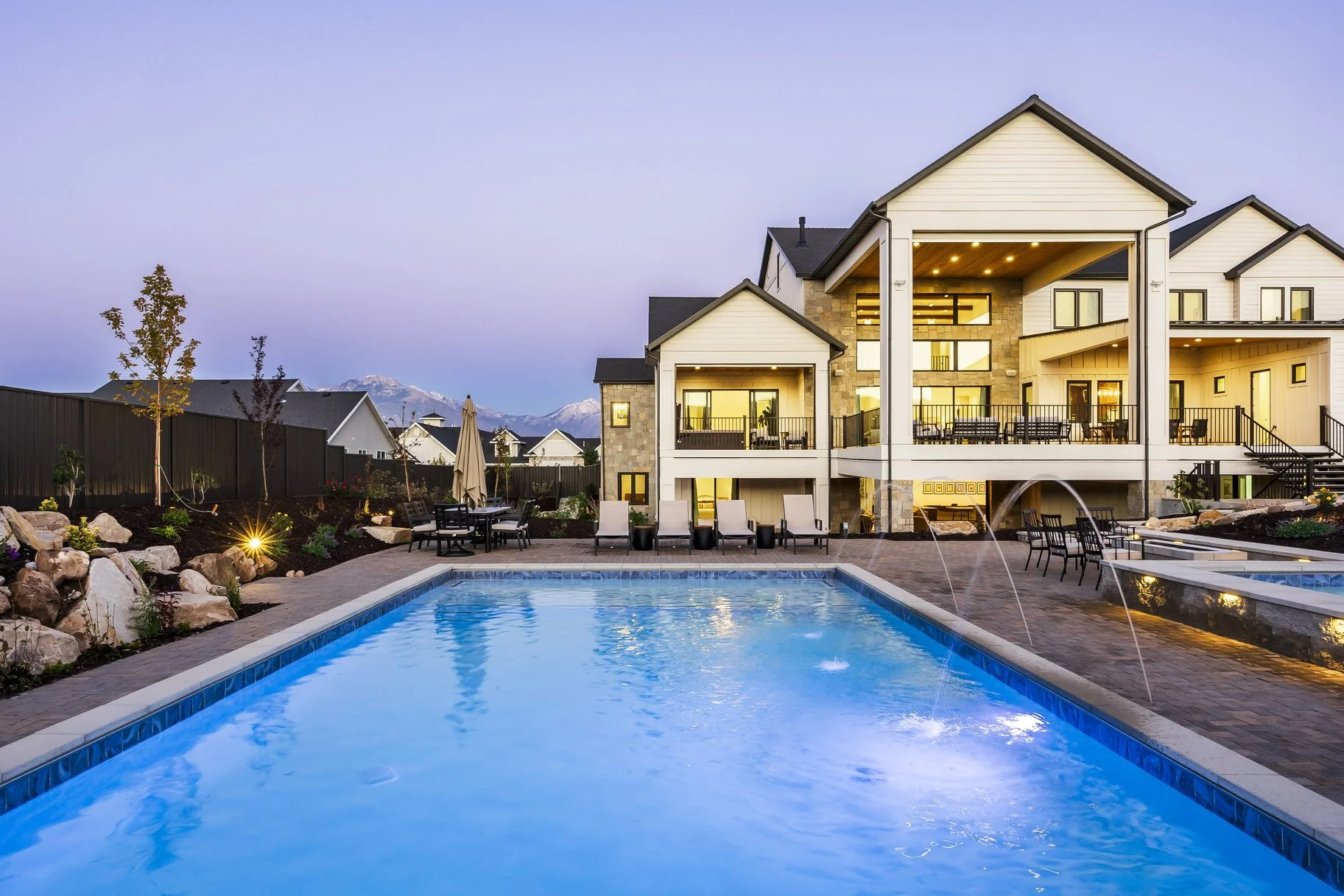 Luxury house with outdoor pool lit up at dusk, surrounded by deck chairs, tables, and garden landscaping, with mountains visible in the background.