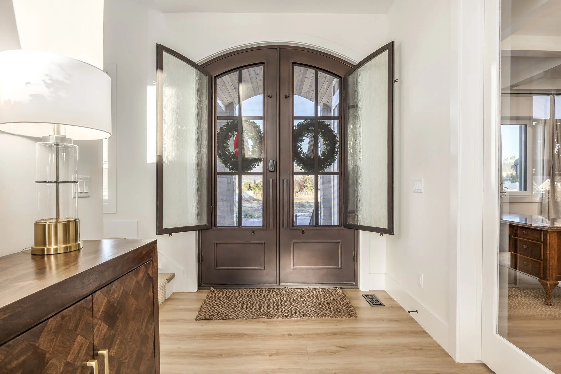 Interior view of a house entryway with wooden double doors with glass panes, wreath decorations, a textured doormat, hardwood flooring, and a side table with a lamp on it.