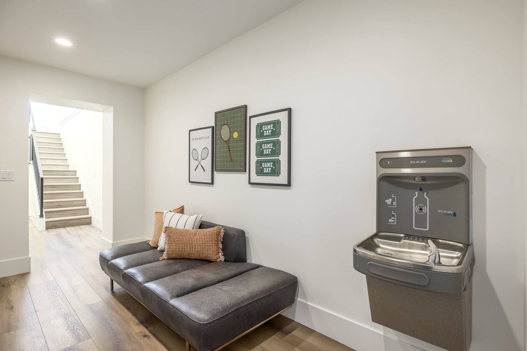Indoor lounge area with a gray sofa, decorative pillows, wall art with tennis theme, and a water fountain on the wall.