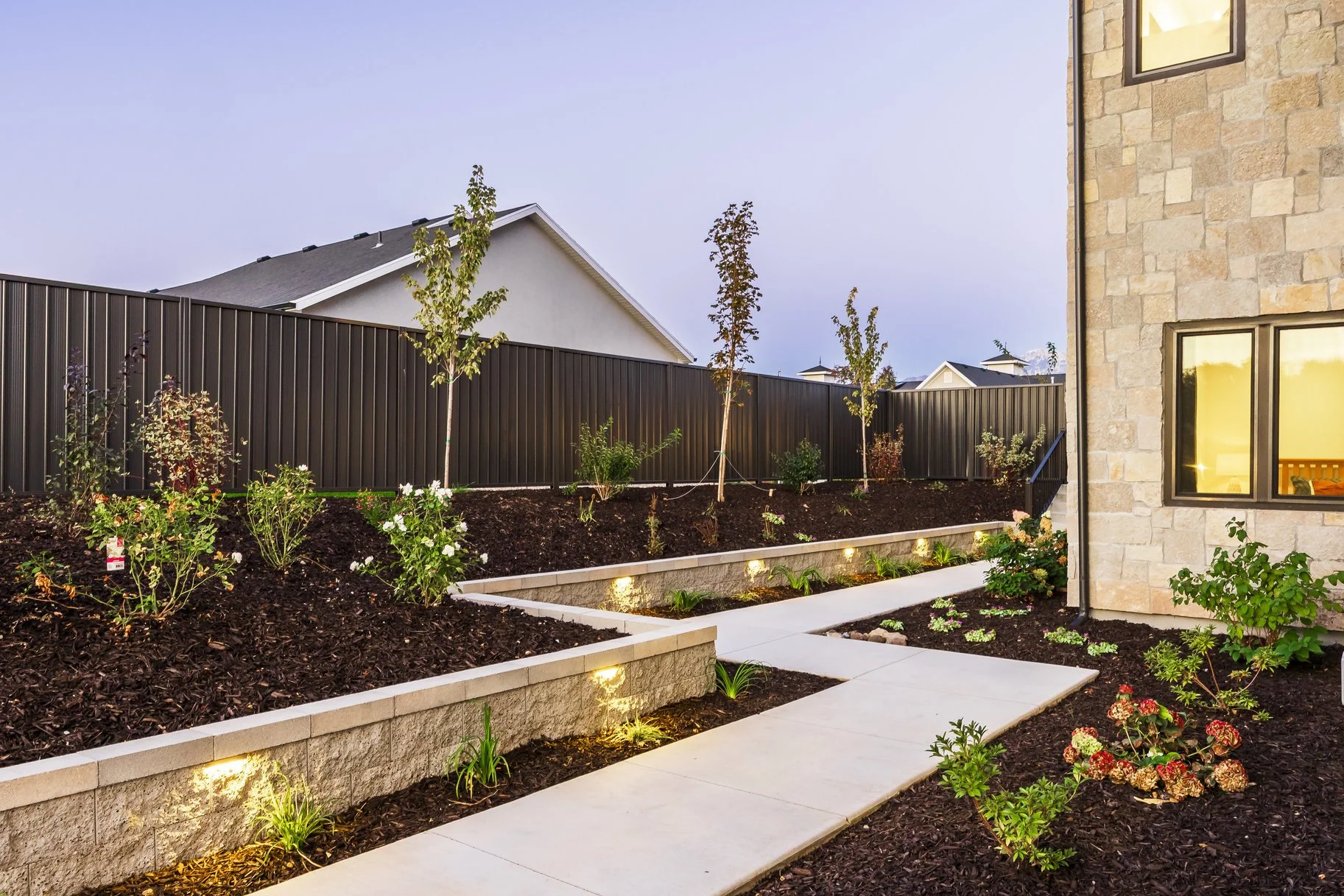  landscaped backyard with newly planted trees and flowers, paved walkway, and exterior of a house with stone siding at dusk.