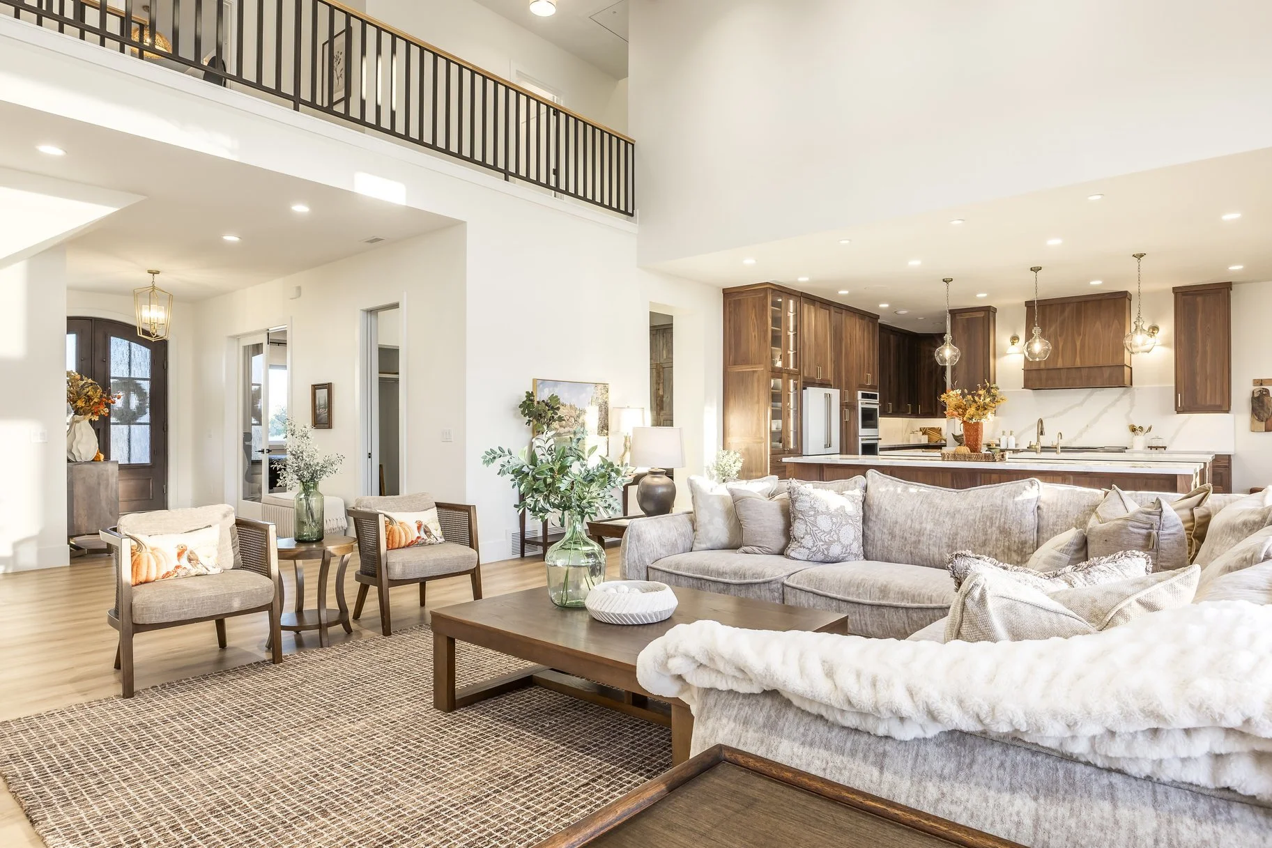 Spacious living room with beige sofas, a wooden coffee table, and decorative pillows. The open kitchen features wooden cabinets, a marble backsplash, and pendant lighting. A front door with a glass panel is visible in the background, along with a bal