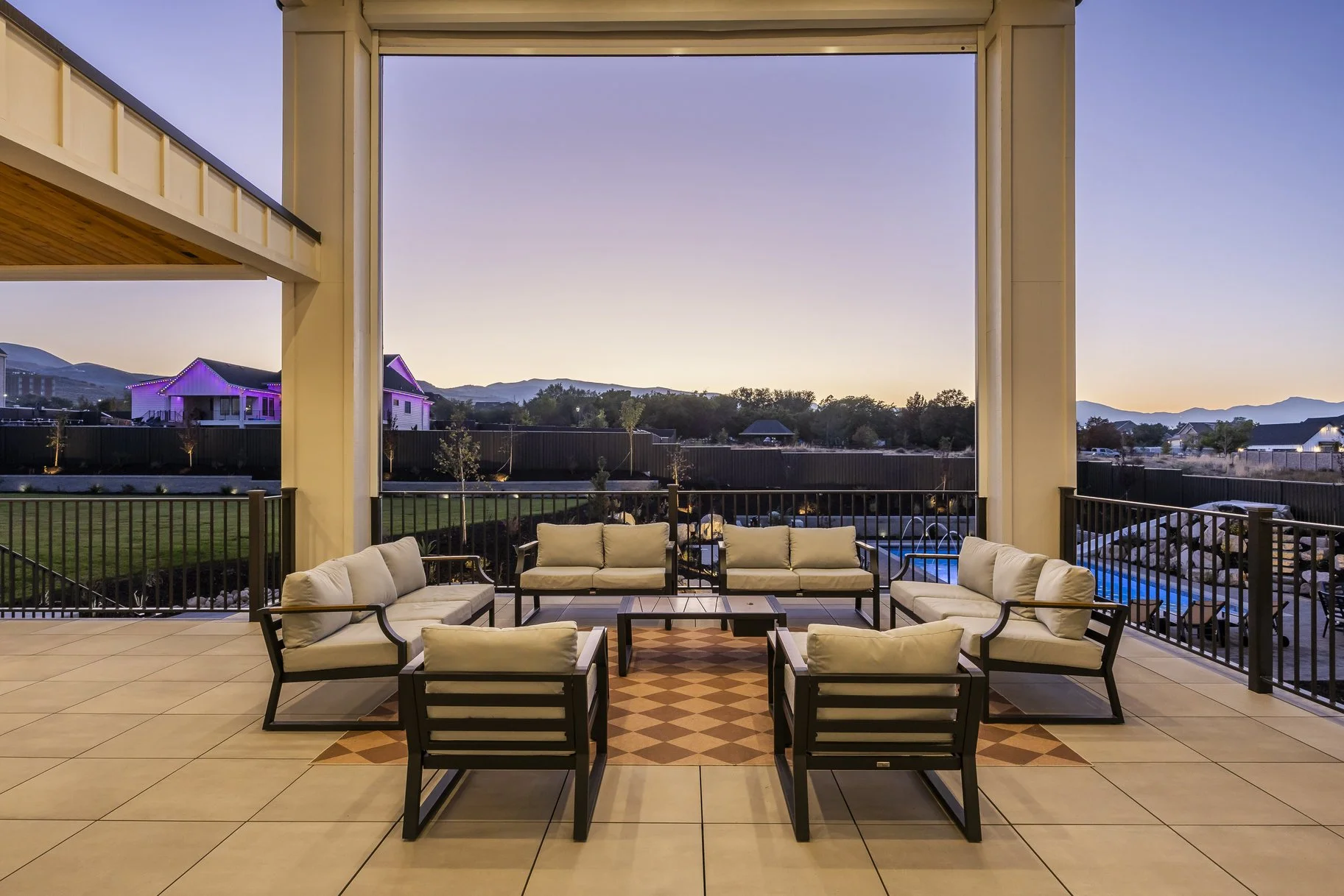 Outdoor patio with beige cushioned seating and a small table, overlooking a fenced yard with a pool and distant houses with mountains in the background during dusk.