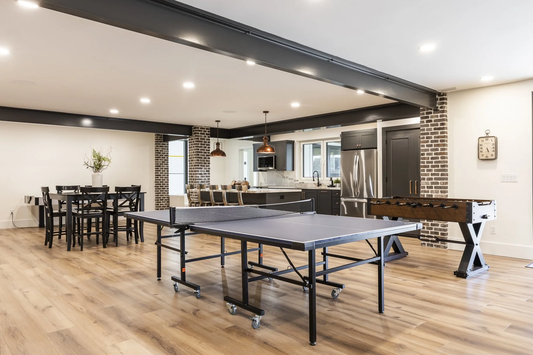 Spacious basement recreation room with ping pong table, foosball table, dining table, and kitchen area with black cabinets and stainless steel appliances.