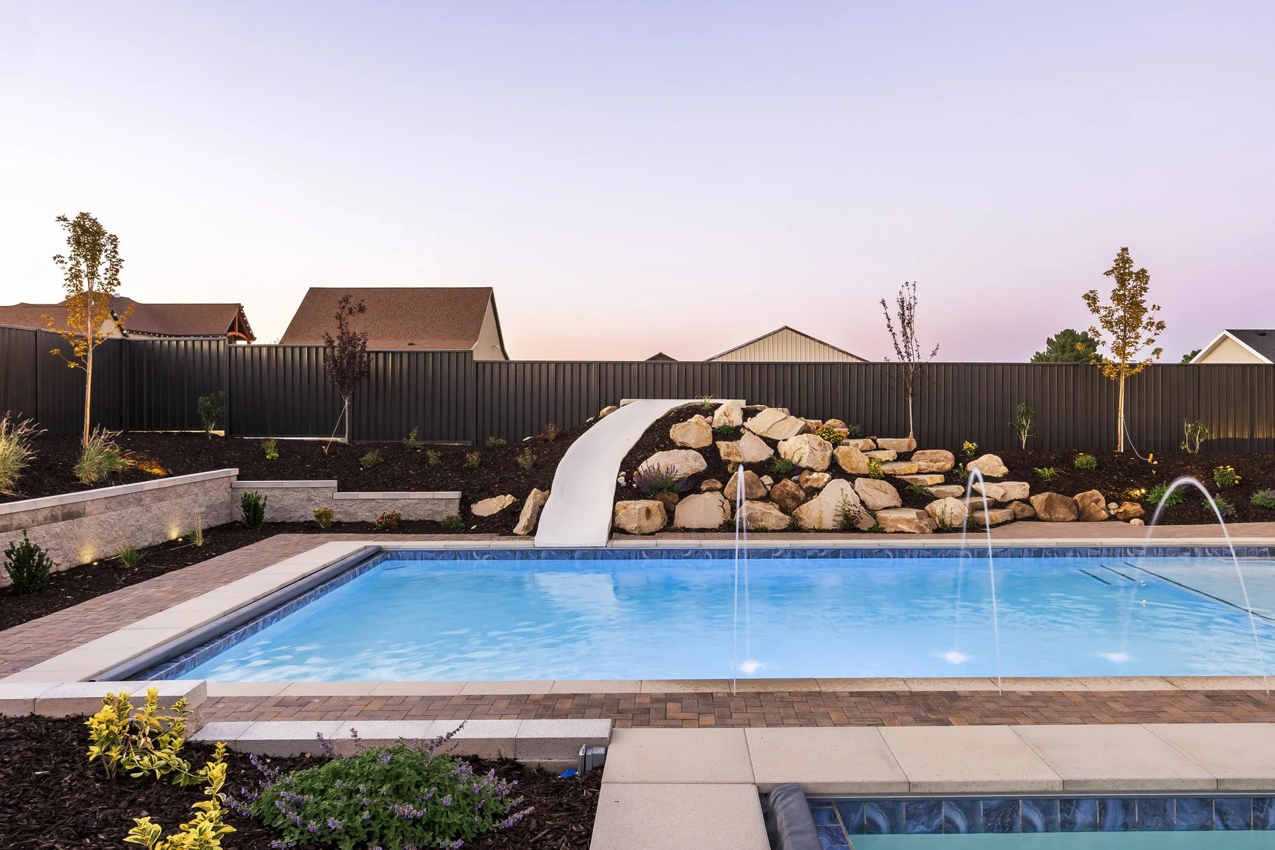 Backyard swimming pool with a waterfall feature and water jets, surrounded by landscaping, small trees, and a dark fence at dusk.