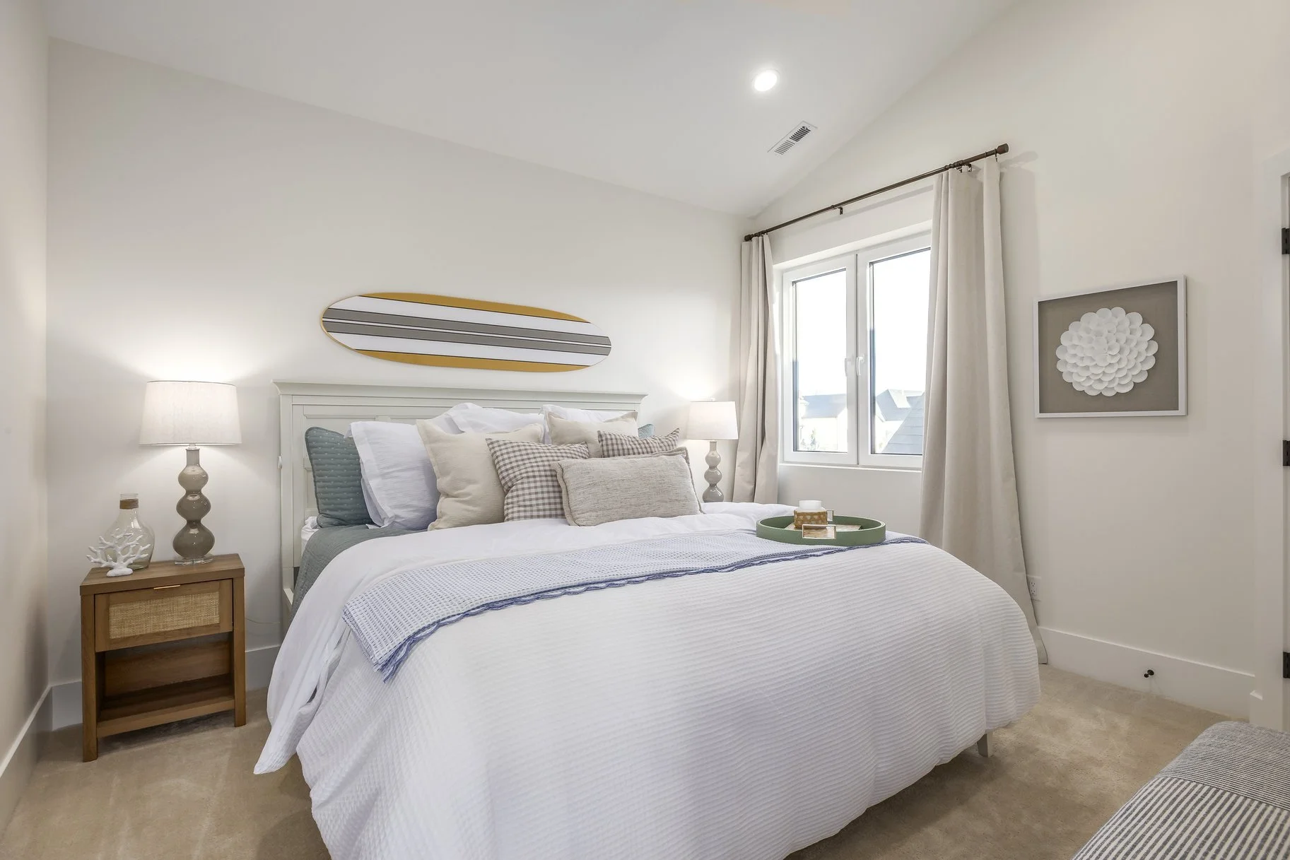 Bedroom with white bedding, multiple pillows, wooden nightstand with lamp, and wall art above the bed. Window with curtains is on the right side.