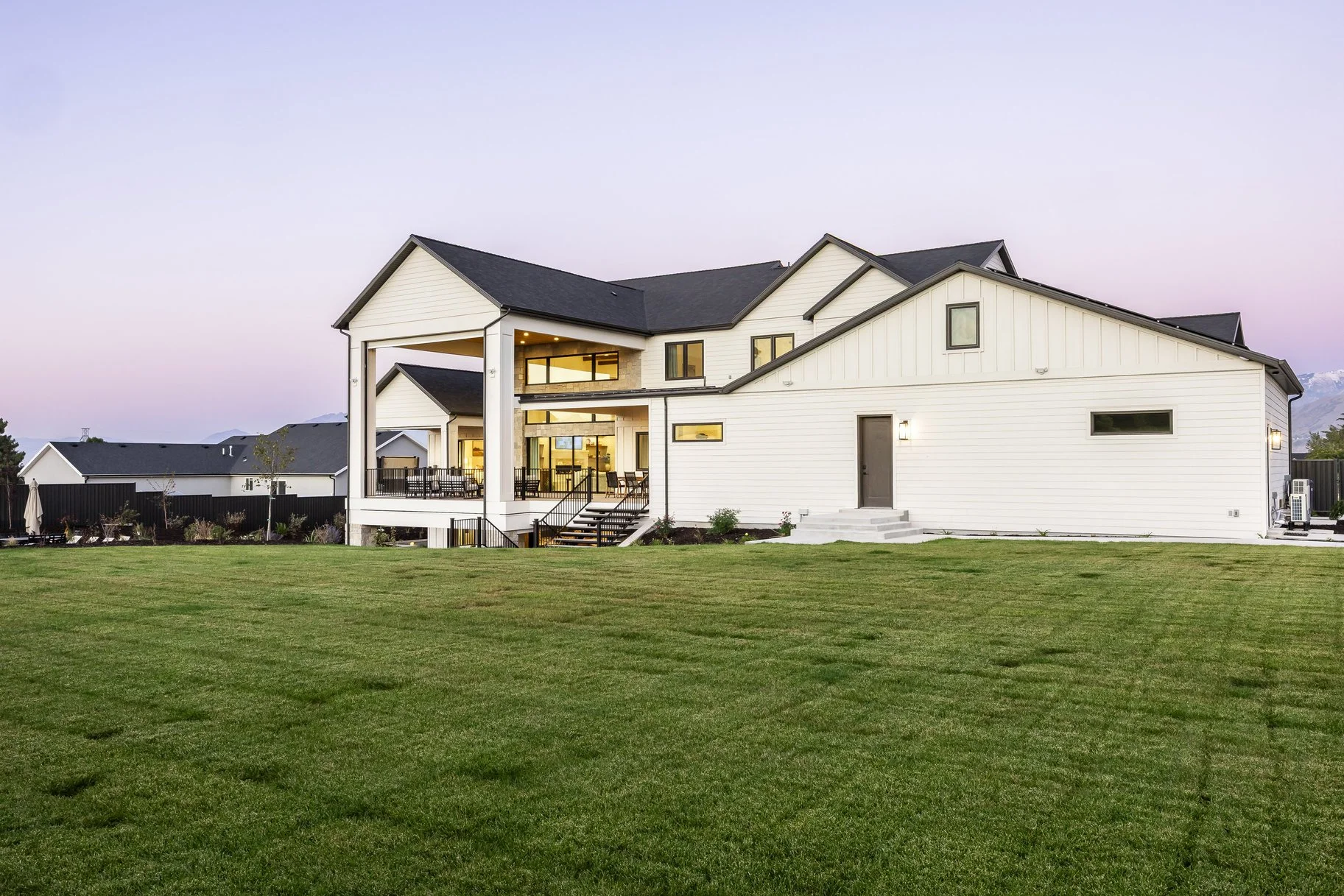 A large modern white house with a black roof, large windows, and a spacious backyard with a green lawn at dusk.