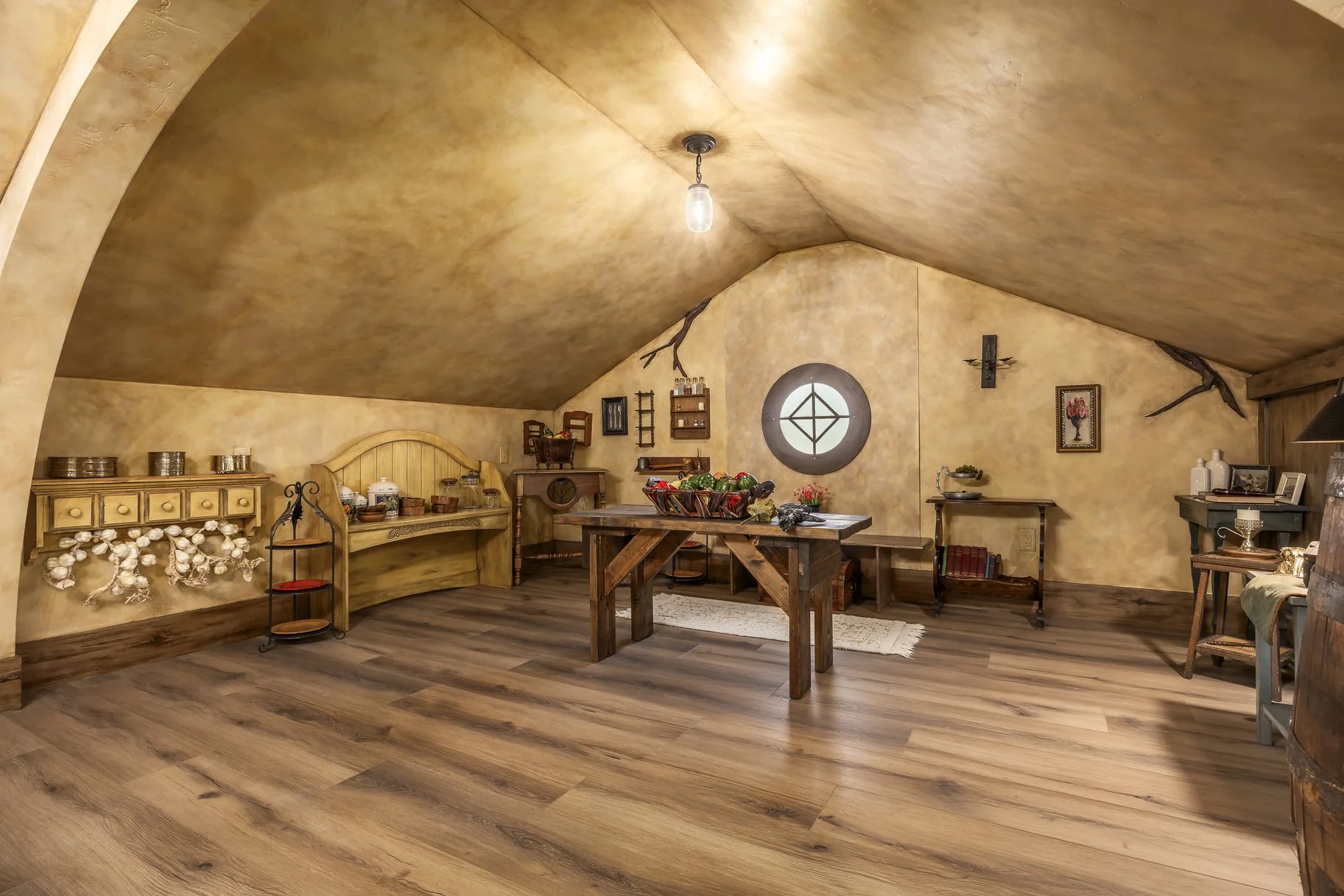 The image shows a cozy attic room with sloped ceiling, decorated in a rustic style with yellowish walls and wooden flooring. There is a wooden table in the center with a basket of vegetables, and various antique and vintage furniture and decorations 