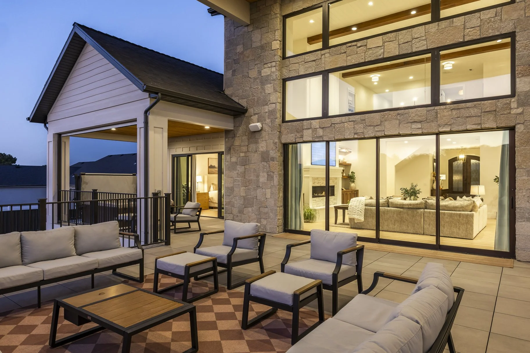 Outdoor patio area with beige cushioned sofas and chairs, a wooden coffee table, and a patterned rug, with a view into a modern living room through large glass sliding doors, on a twilight evening.