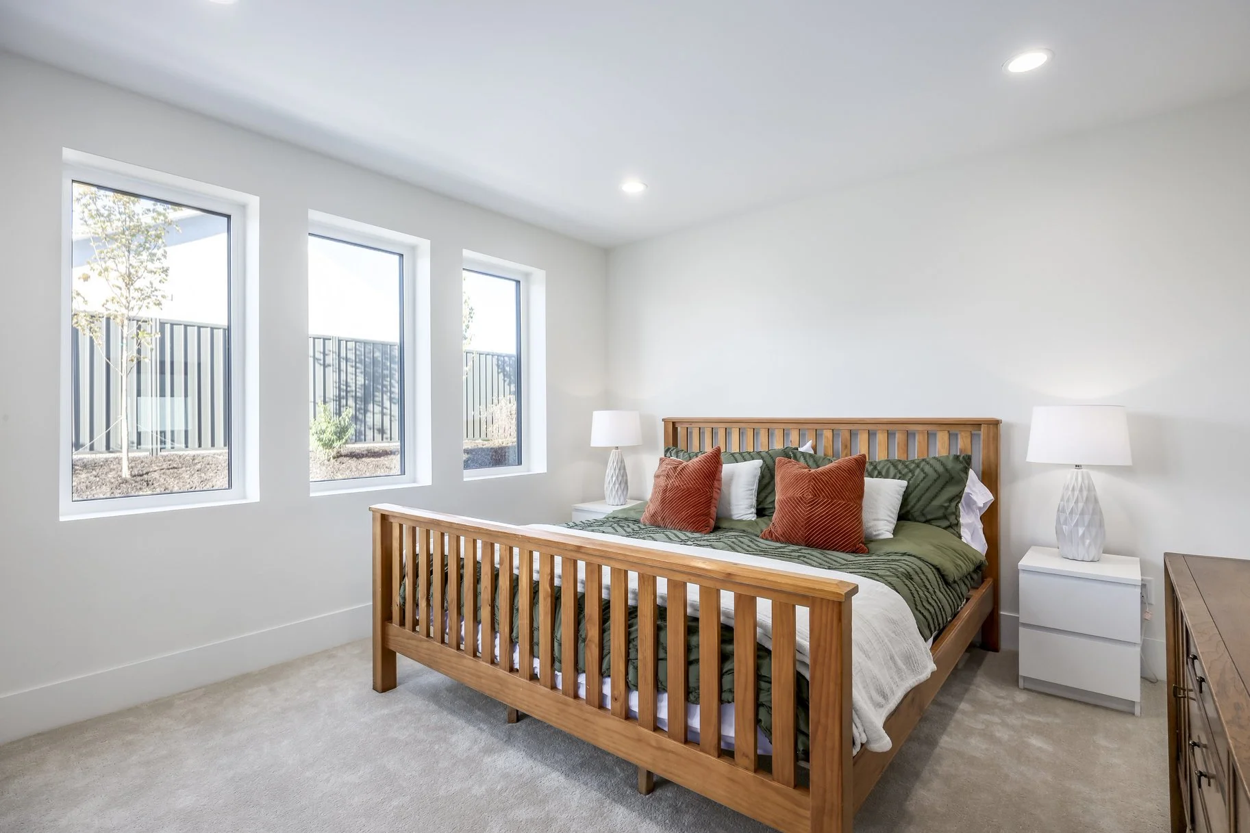 A bedroom with a wooden bed frame, green and white bedding, and decorative pillows. White nightstands with lamps on each side, three large windows, and a beige carpet.